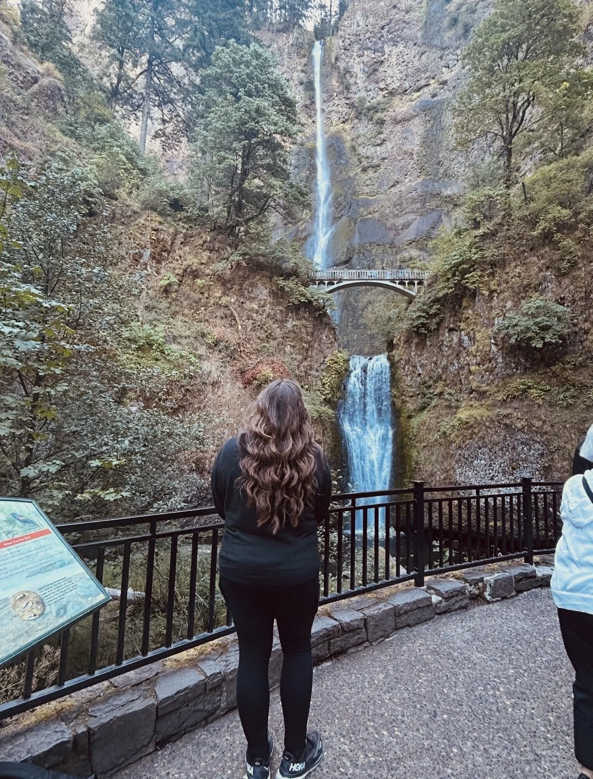 A woman with long curly hair standing at a viewing platform, watching Multnomah Falls with multiple waterfalls cascading down a rocky cliff on a cloudy day.