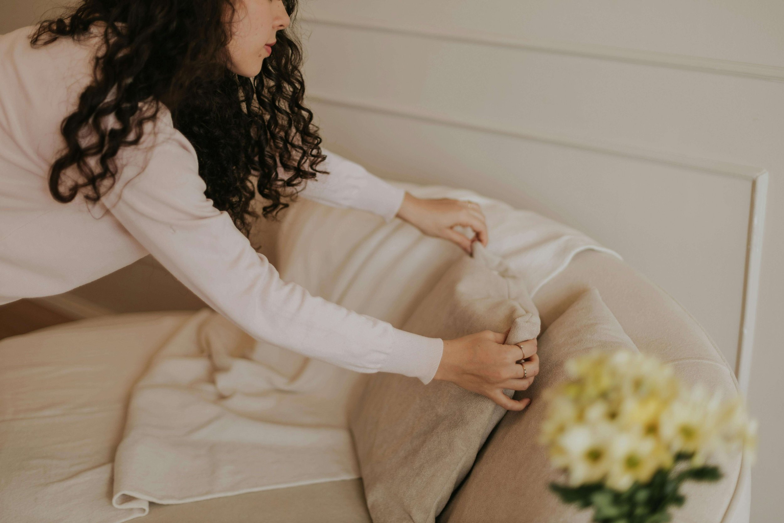 A woman with curly dark hair making the bed with beige pillows on a cream-colored couch.