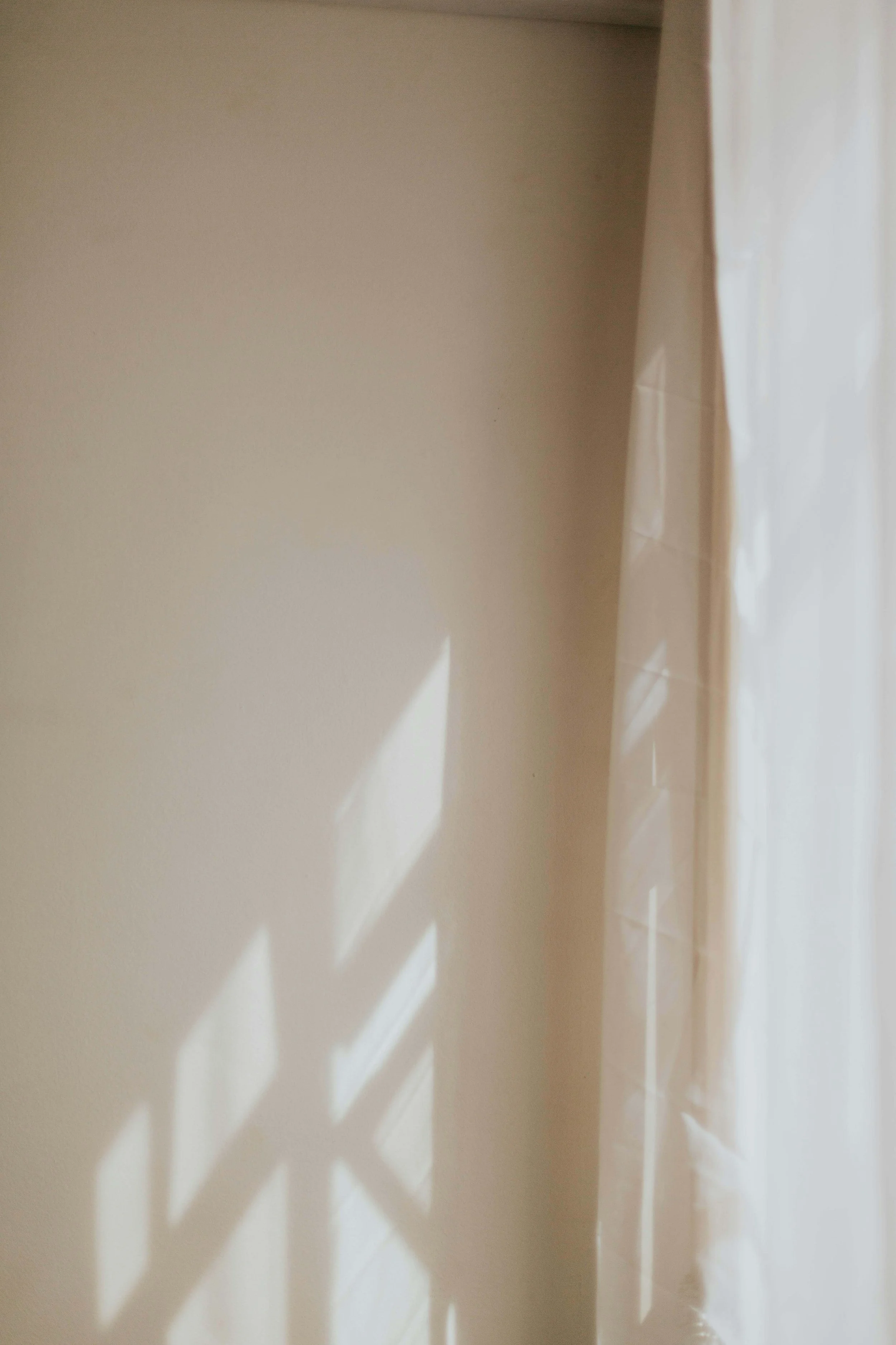 Sunlight filtering through window blinds casting shadows on a beige wall near white curtains.
