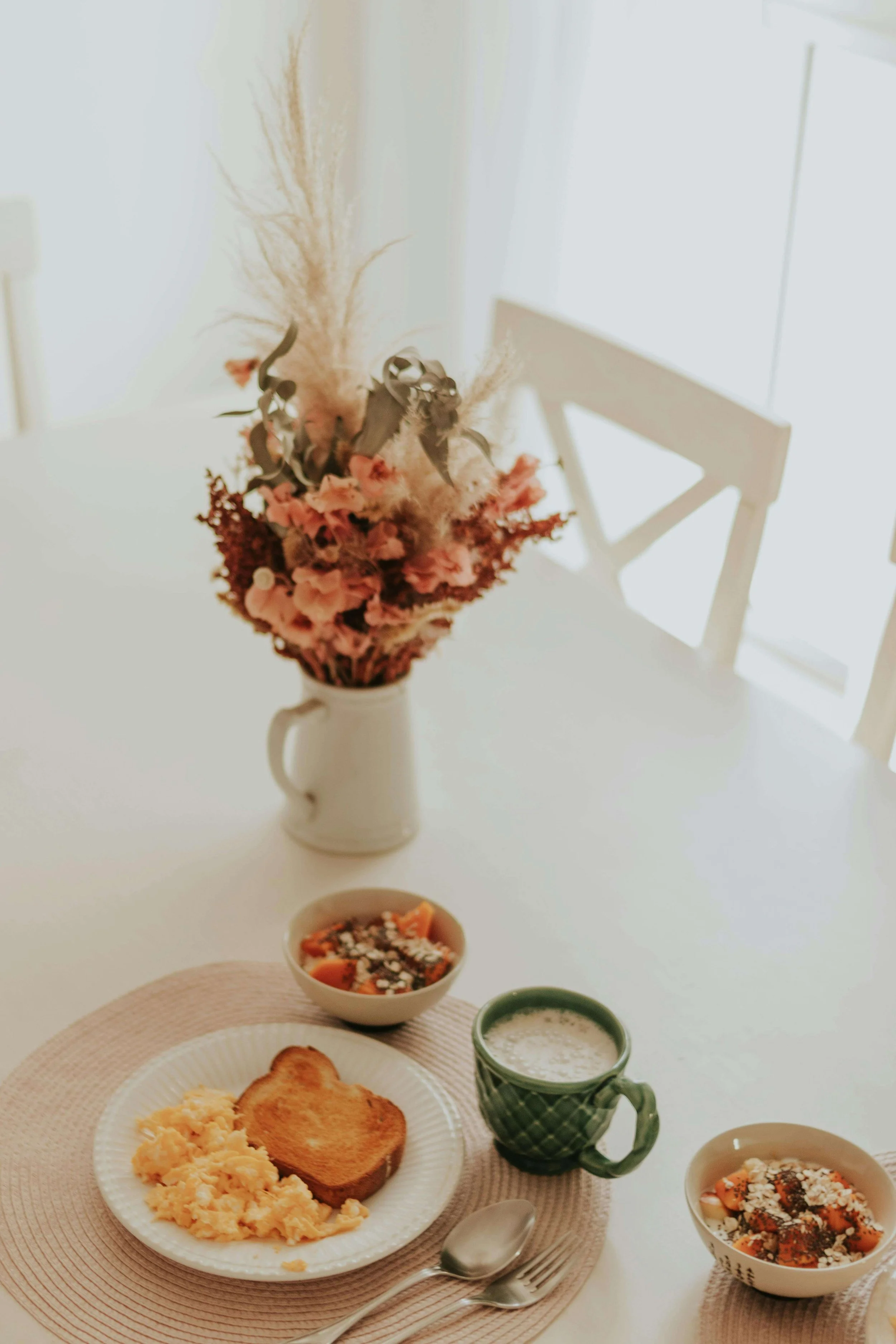 A breakfast table with scrambled eggs, toasted bread, a bowl of fruit, and a mug of milk. There's a vase with pink and beige dried flowers and plants as a centerpiece.