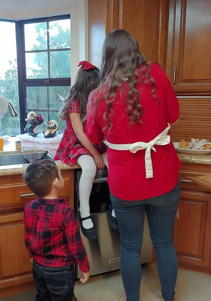 A woman with long curly hair dressed in a red top and blue jeans is helping a young girl sitting on the kitchen counter with preparing the turkey, while a boy stands nearby watching. The kitchen has wooden cabinets and a window with a view outside.