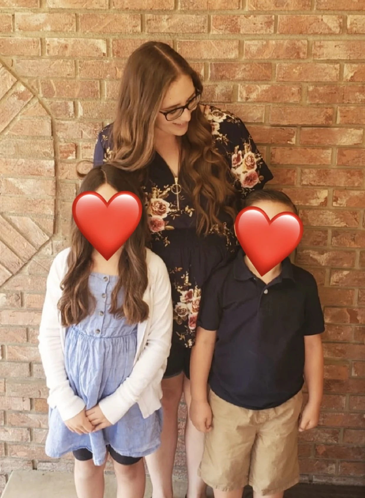 A woman with long brown hair and glasses is standing behind two children, a girl and a boy, against a brick wall. The children's faces are obscured by large red hearts. The girl has long wavy hair, is wearing a light blue dress with buttons and a white cardigan, and is standing with her hands clasped. The boy has short hair, is wearing a navy polo shirt and khaki shorts, and stands with his right arm hanging loosely by his side.