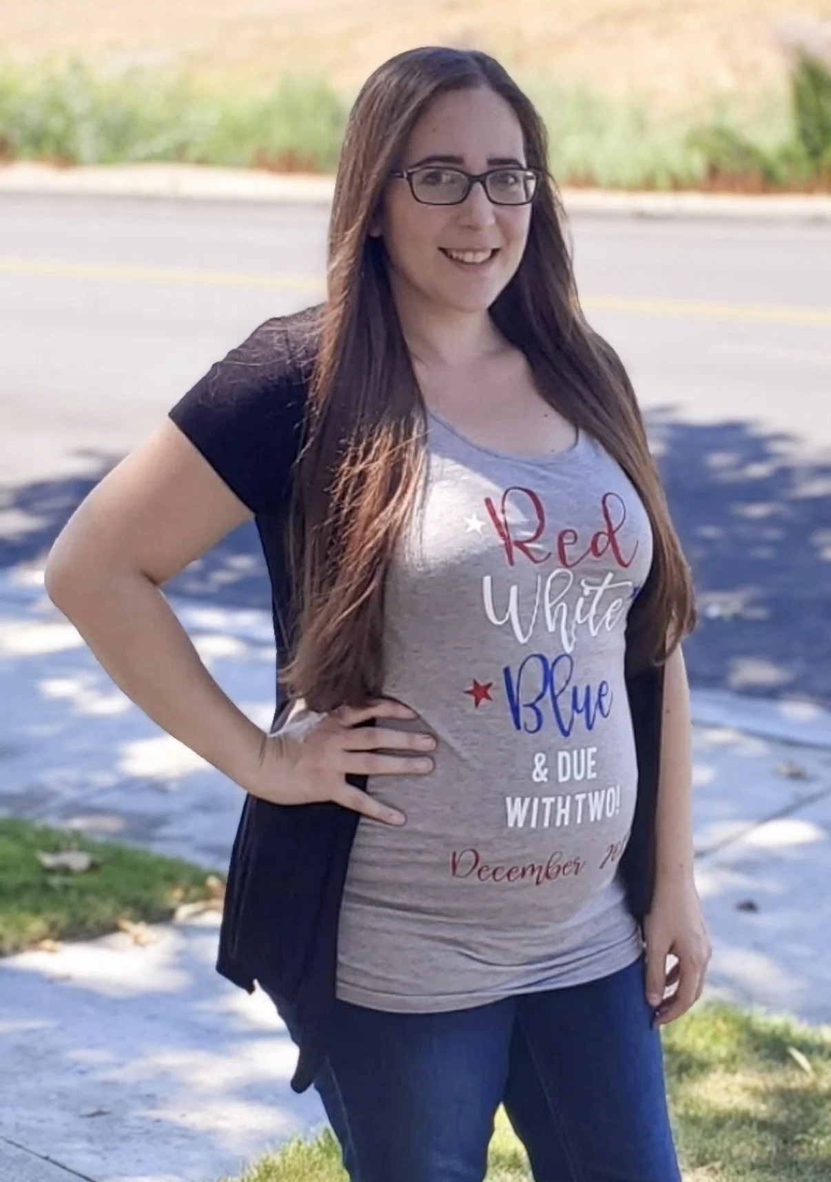 A young woman with long brown hair, glasses, and a black and gray T-shirt that reads "Red White & Blue & Due With Two! December" stands outdoors with her hand on her hip, smiling at the camera. The background features a sidewalk, grass, trees, and a street.