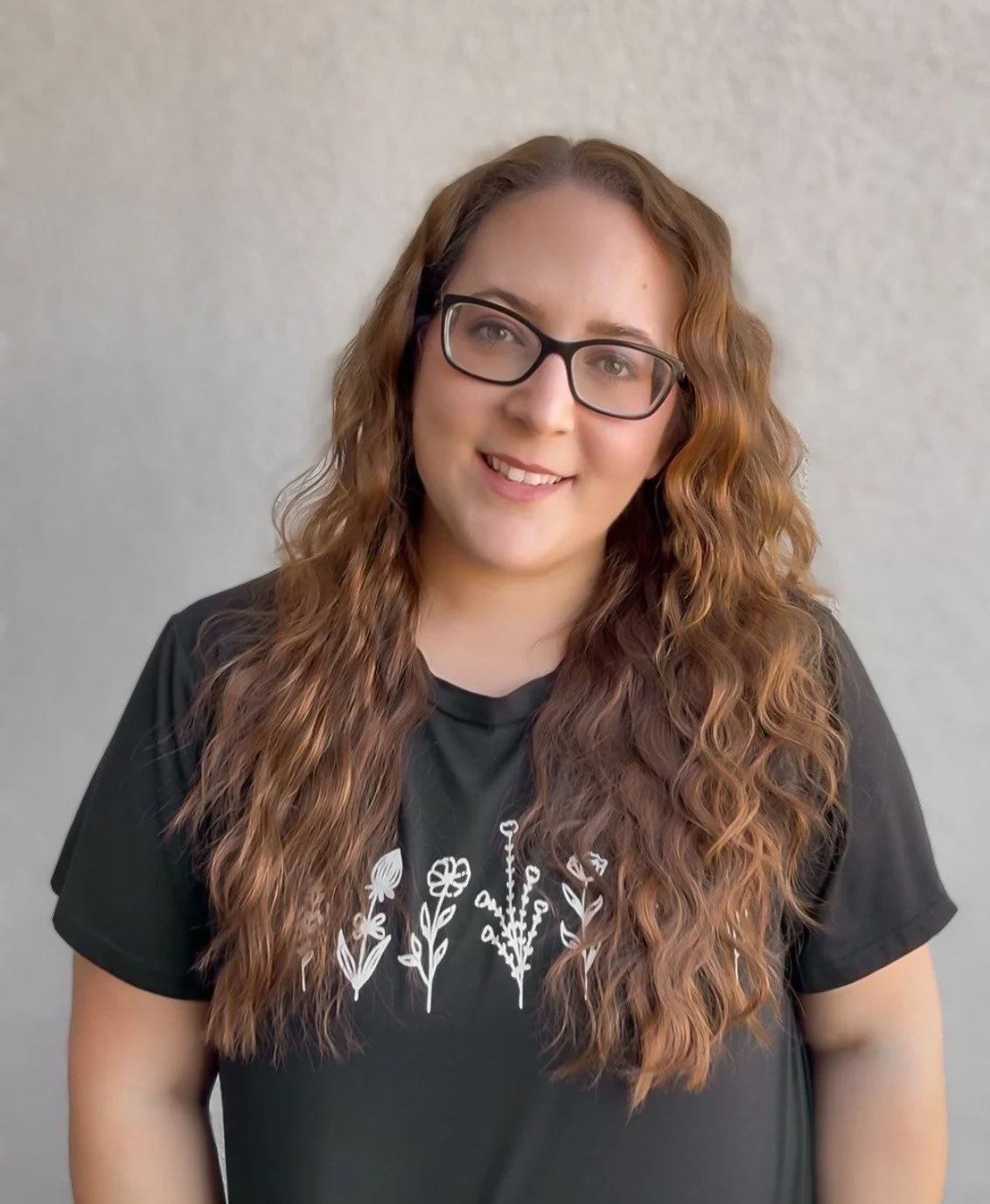 A young woman with long, wavy red hair, wearing glasses and a black T-shirt with white floral embroidery, smiling at the camera.