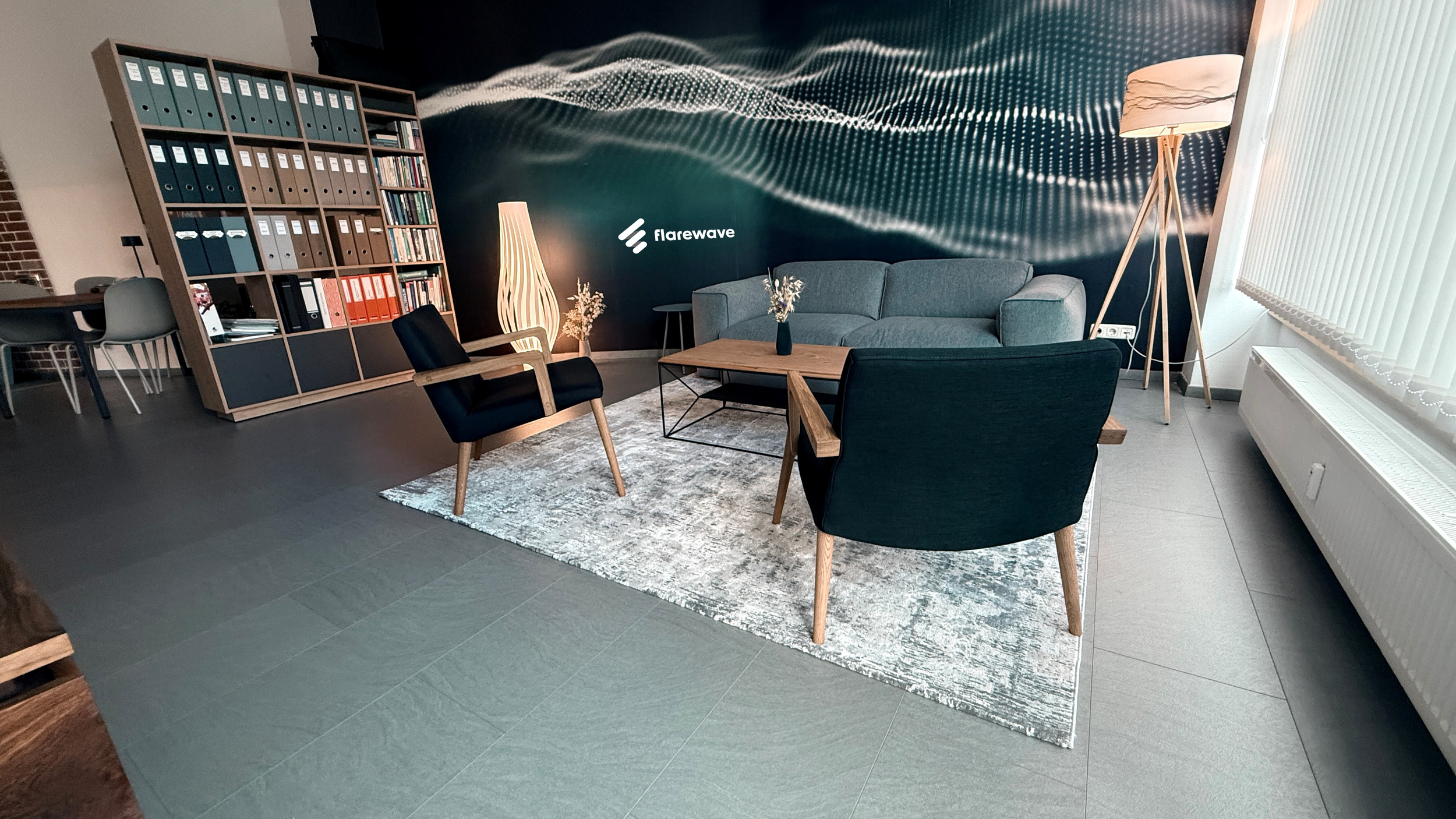 Modernes Wohnzimmer mit grauem Sofa, zwei schwarzen Sessel, einem Holztisch, Bücherregal und Stehlampen, Wand mit digitalem Wellenmuster und dem Logo 'flarewave'