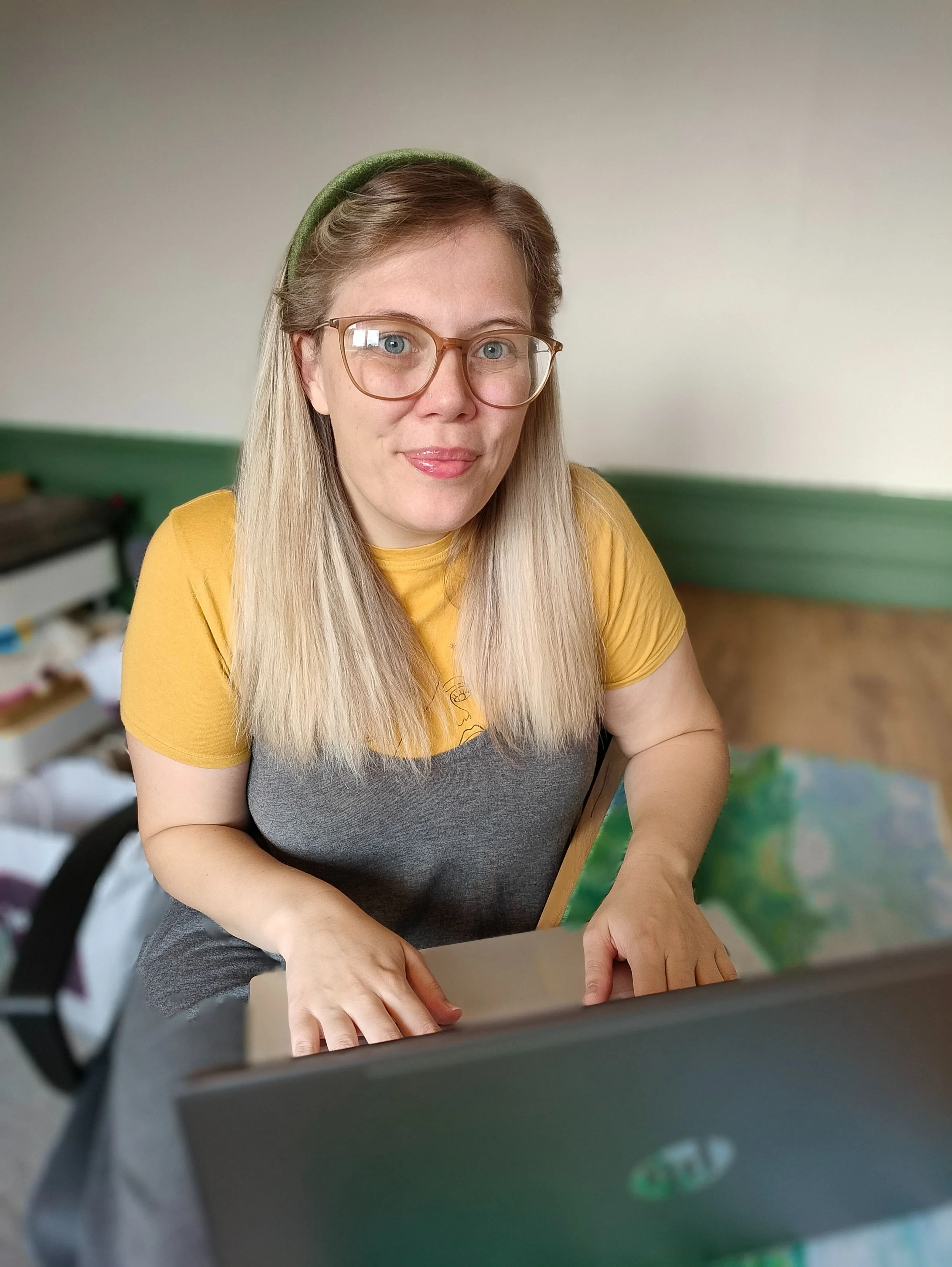 A woman with blonde hair, wearing glasses, a green headband, a yellow shirt, and a gray apron, sitting at a table with her hands on a laptop keyboard.