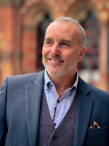 A smiling middle-aged man with a beard and short gray hair, wearing a blue blazer over a blue shirt and a dark vest, standing outdoors with a blurred brick building in the background.