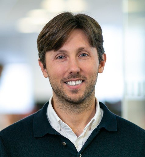 A smiling man with medium-length brown hair, wearing a white collared shirt and a dark cardigan, standing in a bright indoor setting.