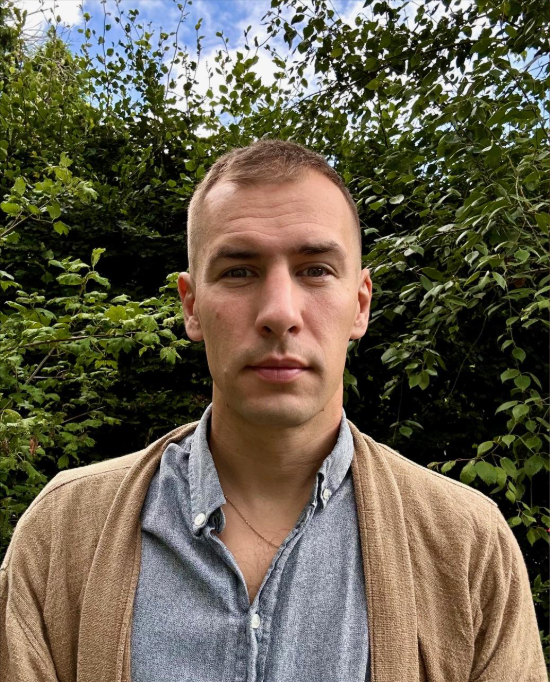 A young man standing outdoors in front of green bushes and trees, wearing a light gray button-up shirt and a tan cardigan, with short light brown hair and a neutral expression.