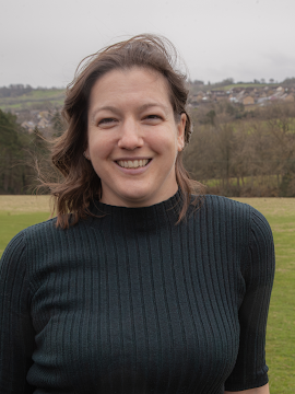 A woman with shoulder-length brown hair smiling outdoors in a grassy field with hills and houses in the background.
