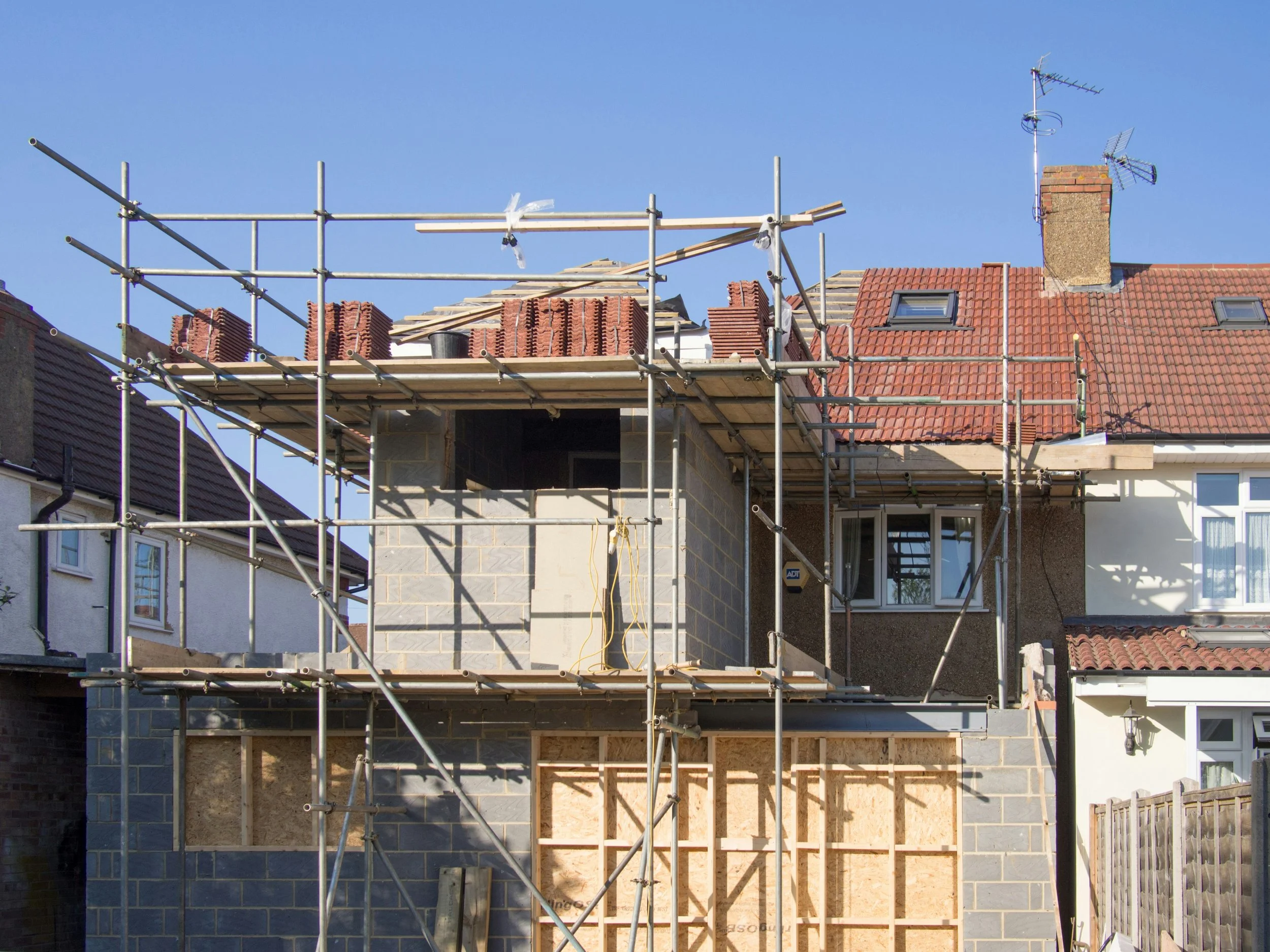 House under construction with scaffolding and brickwork, and a neighboring house with a red-tiled roof and satellite dishes.