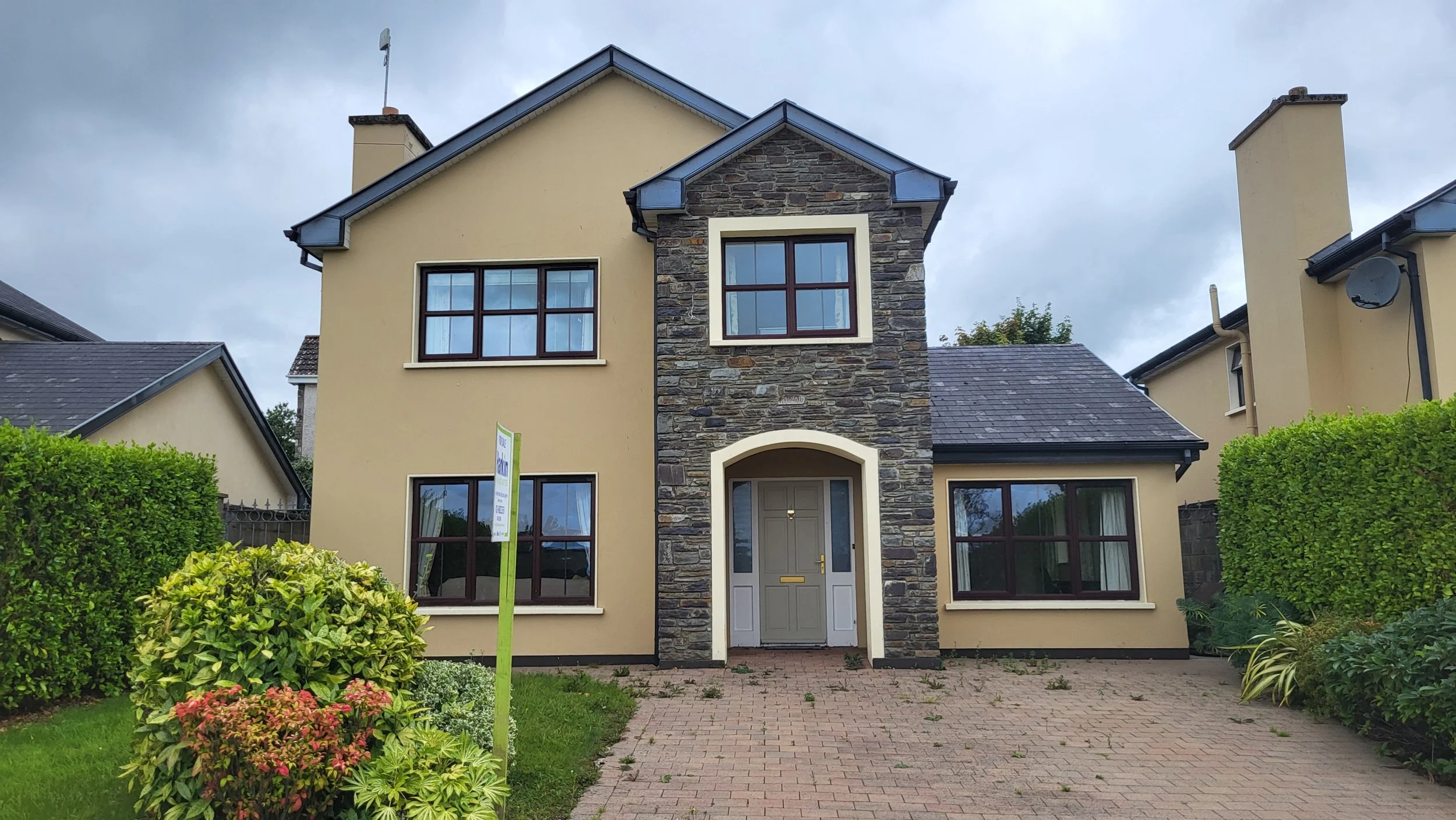 Front view of a two-story house with yellow exterior walls, a stone section around the entrance, and dark brown window frames. The house has a paved driveway and green landscaping with plants and bushes.