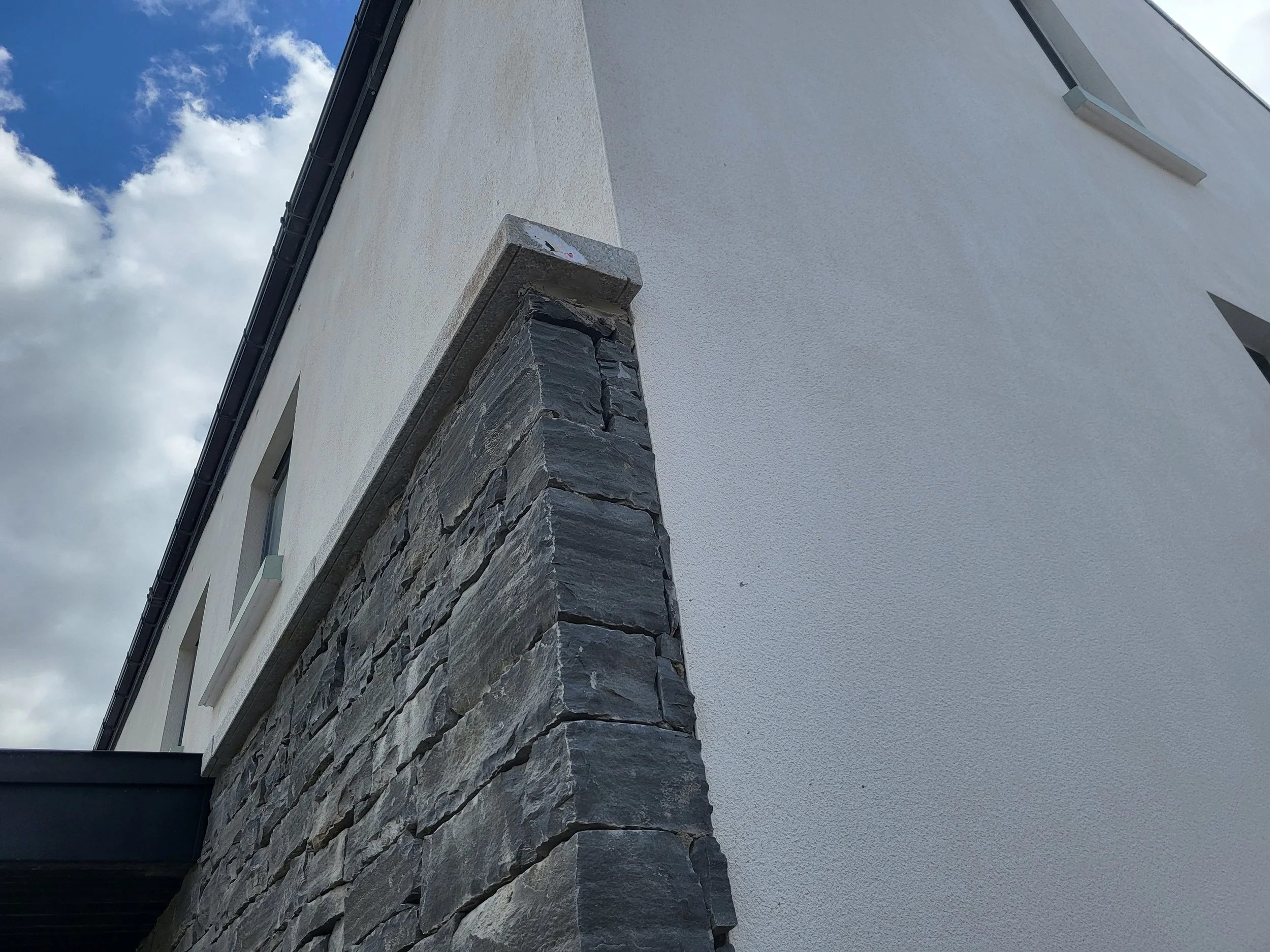 Close-up of the corner of a modern building, showing a white stucco wall, a stacked slate stone corner, and a gray concrete cap, against a background of a partly cloudy sky.