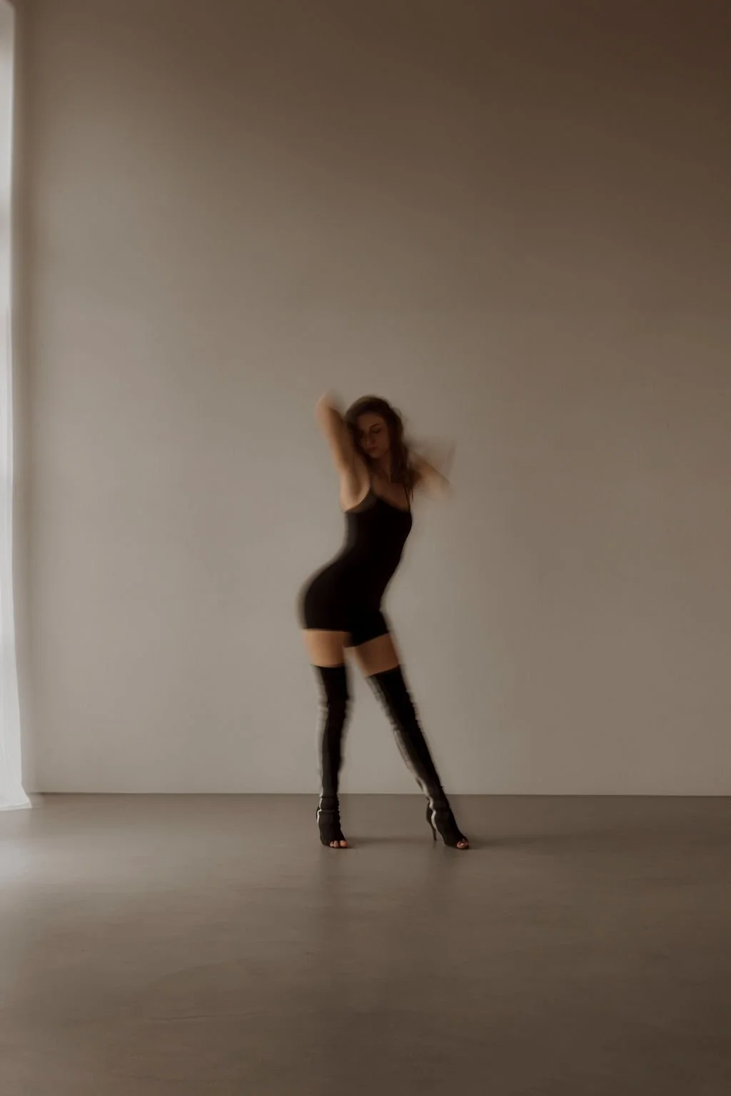 A woman standing in a minimalist room with a plain wall and hardwood floor, wearing a black dress and thigh-high black boots, striking a pose with her arms raised behind her head.