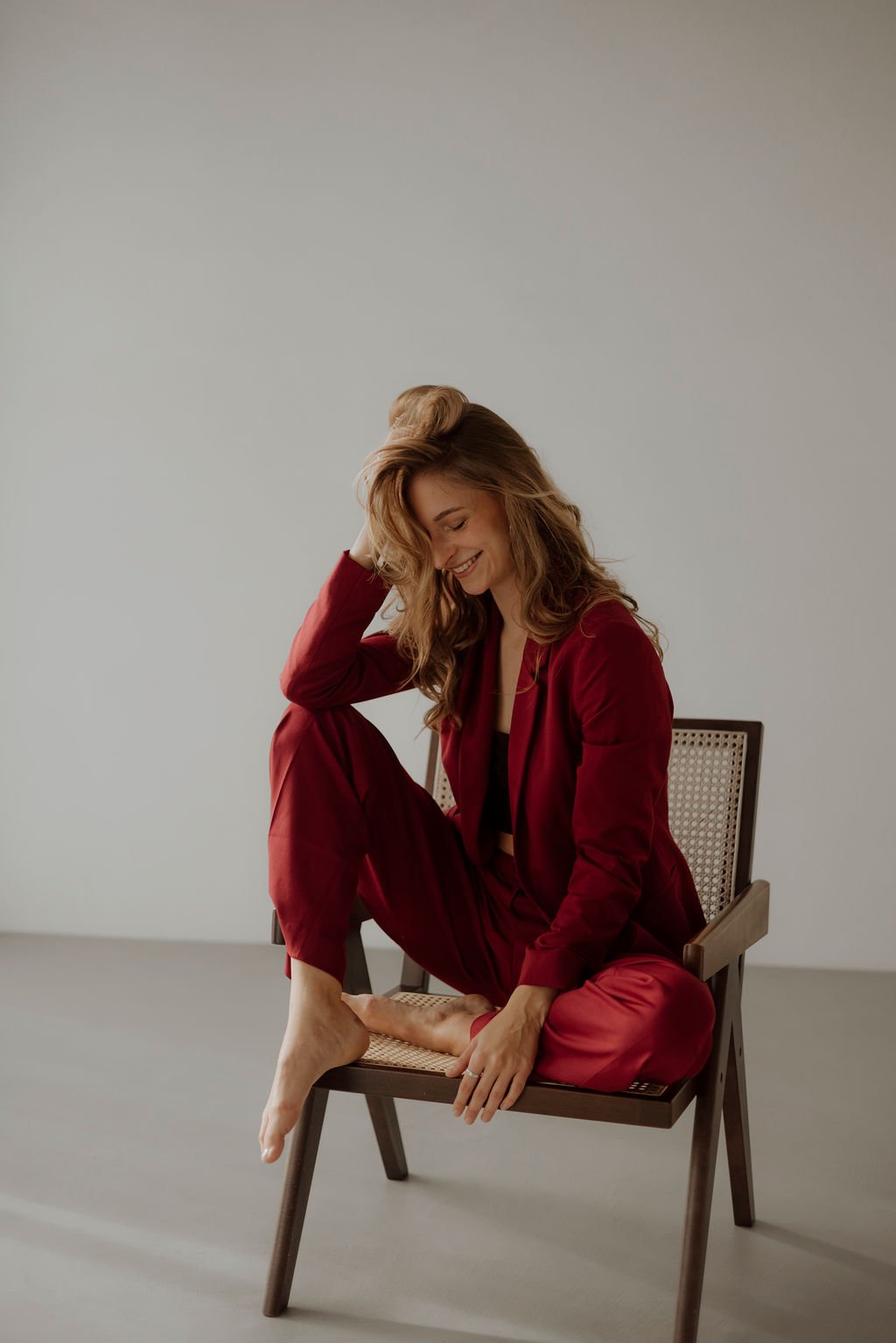 Junge Frau in rotem Anzug sitzt auf einem Stuhl, lächelt und streicht sich durch die Haare, im minimalistischen Raum.