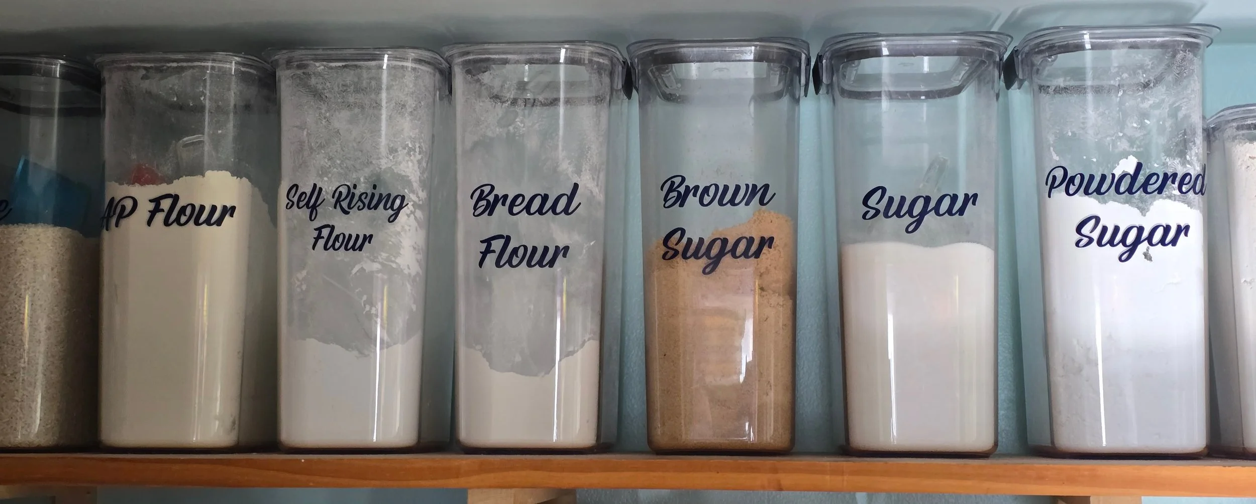Pantry shelf with clear containers labeled 'All Purpose Flour,' 'Self Rising Flour,' 'Bread Flour,' 'Brown Sugar,' 'Sugar,' and 'Powdered Sugar' filled with different types of flour and sugar.
