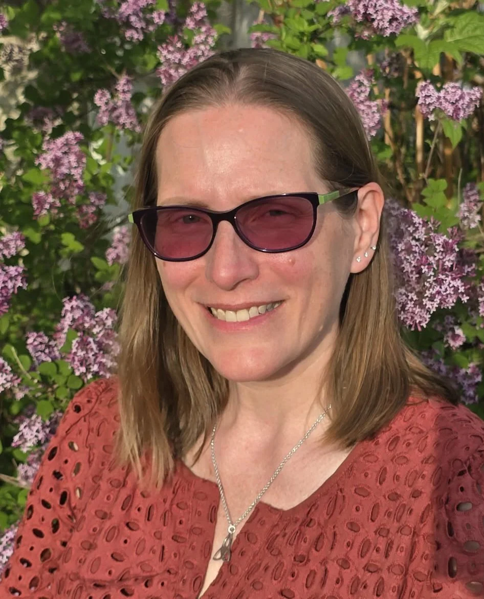 A woman wearing sunglasses and a reddish top standing in front of pink and purple flowers and green foliage, smiling at the camera.