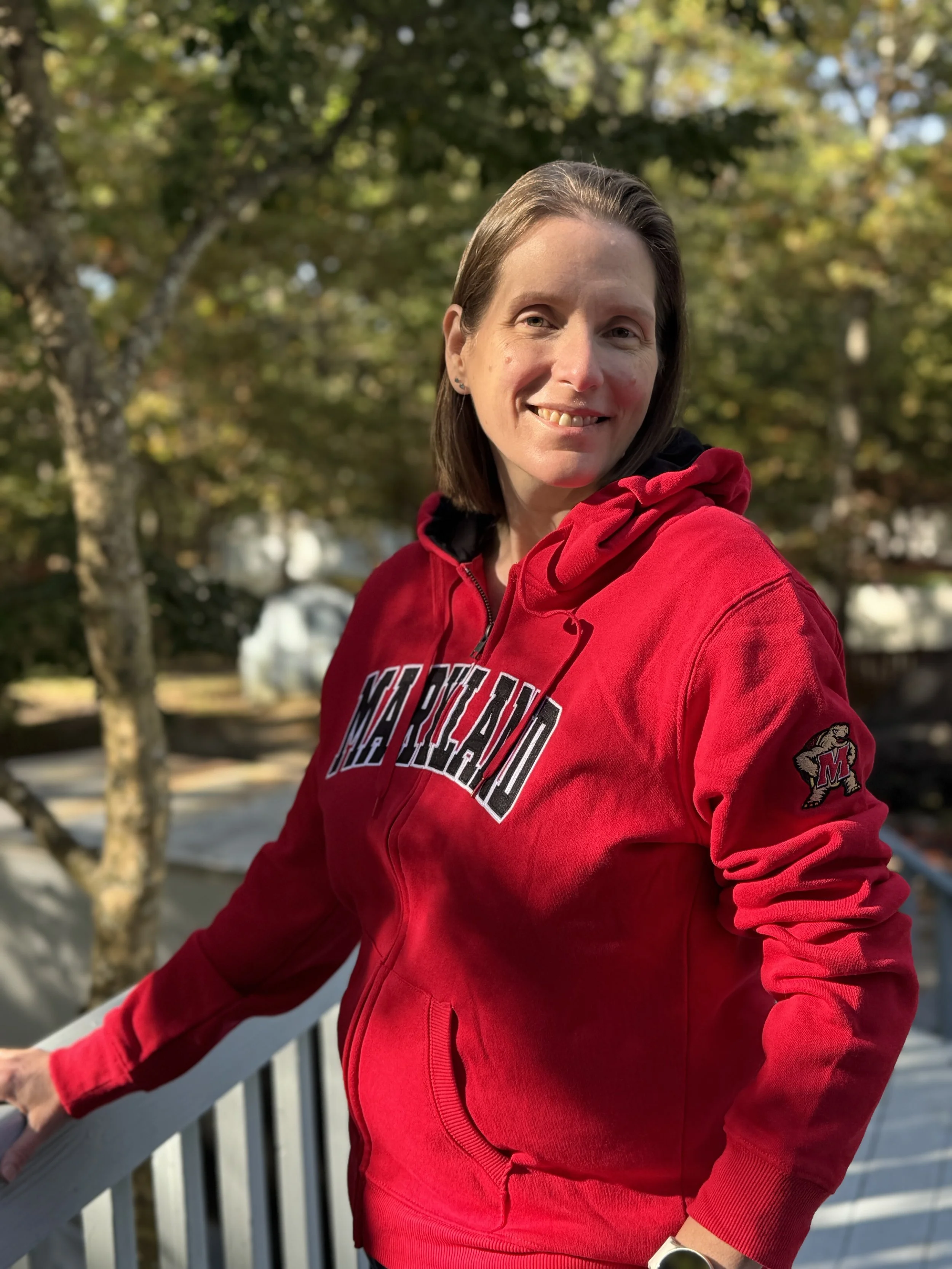 A woman with shoulder-length brown hair, smiling, wearing a red Maryland hoodie, standing outdoors on a balcony with trees and houses in the background during fall.