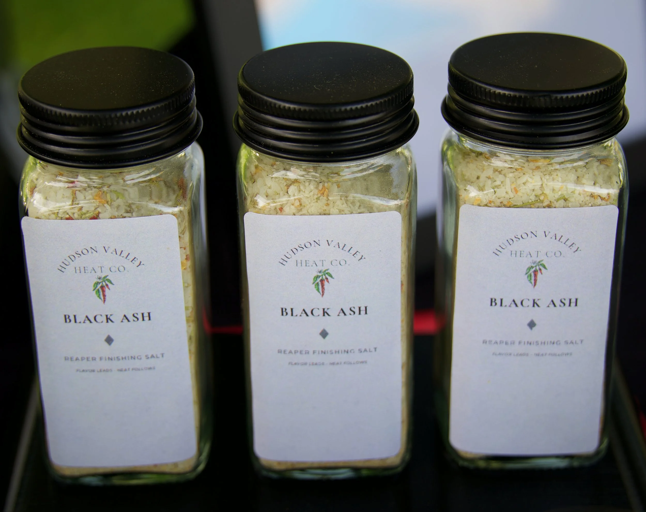 Three glass jars with black screw-on lids containing finishing salt, labeled 'Black Ash' from Hudson Valley Heat Co.