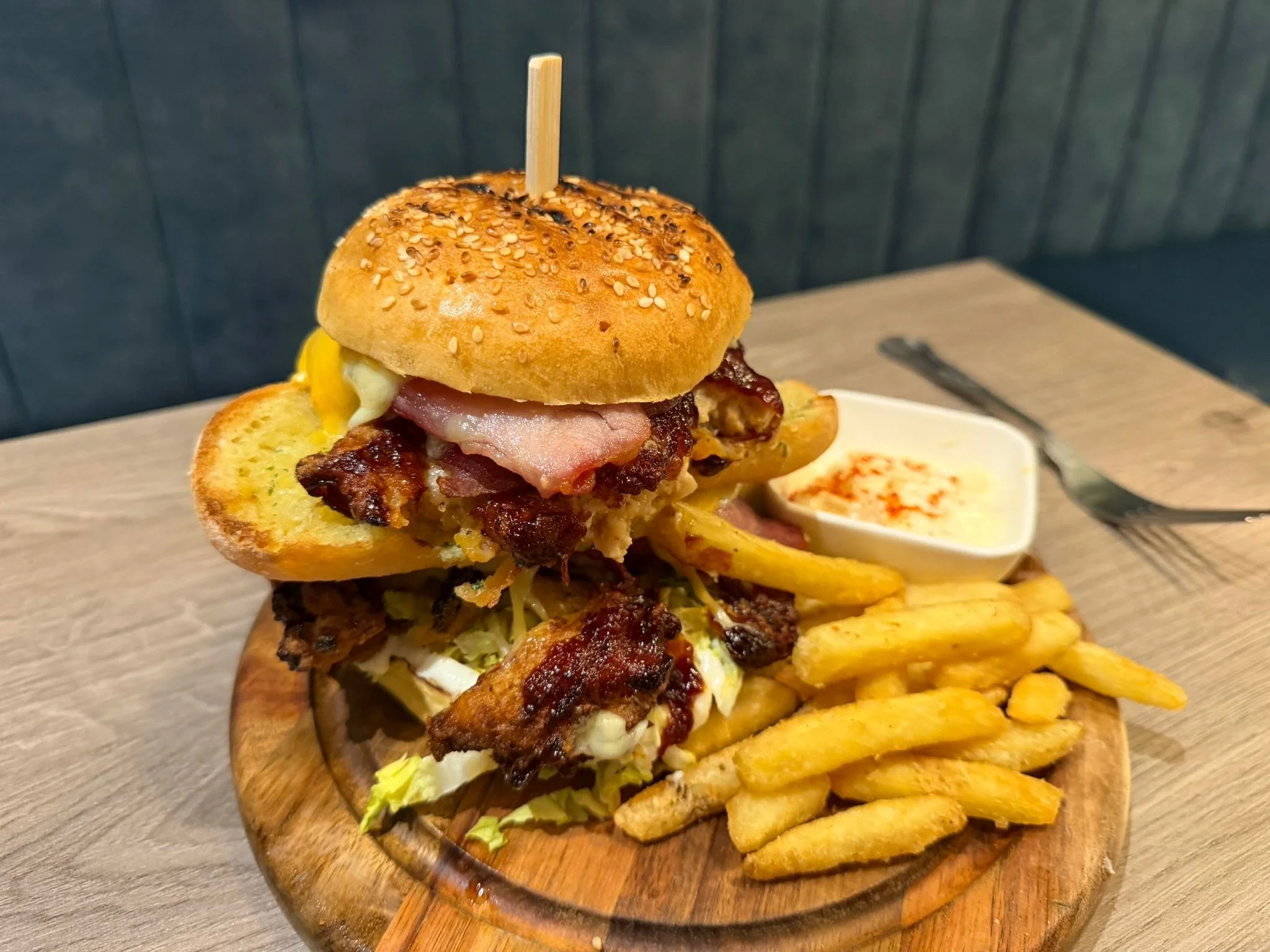 A towering burger on a wooden serving board with French fries, topped with onion rings, cheese, lettuce, tomato, and a sausage link, with a chocolate stick protruding from the onion rings, in a restaurant setting.