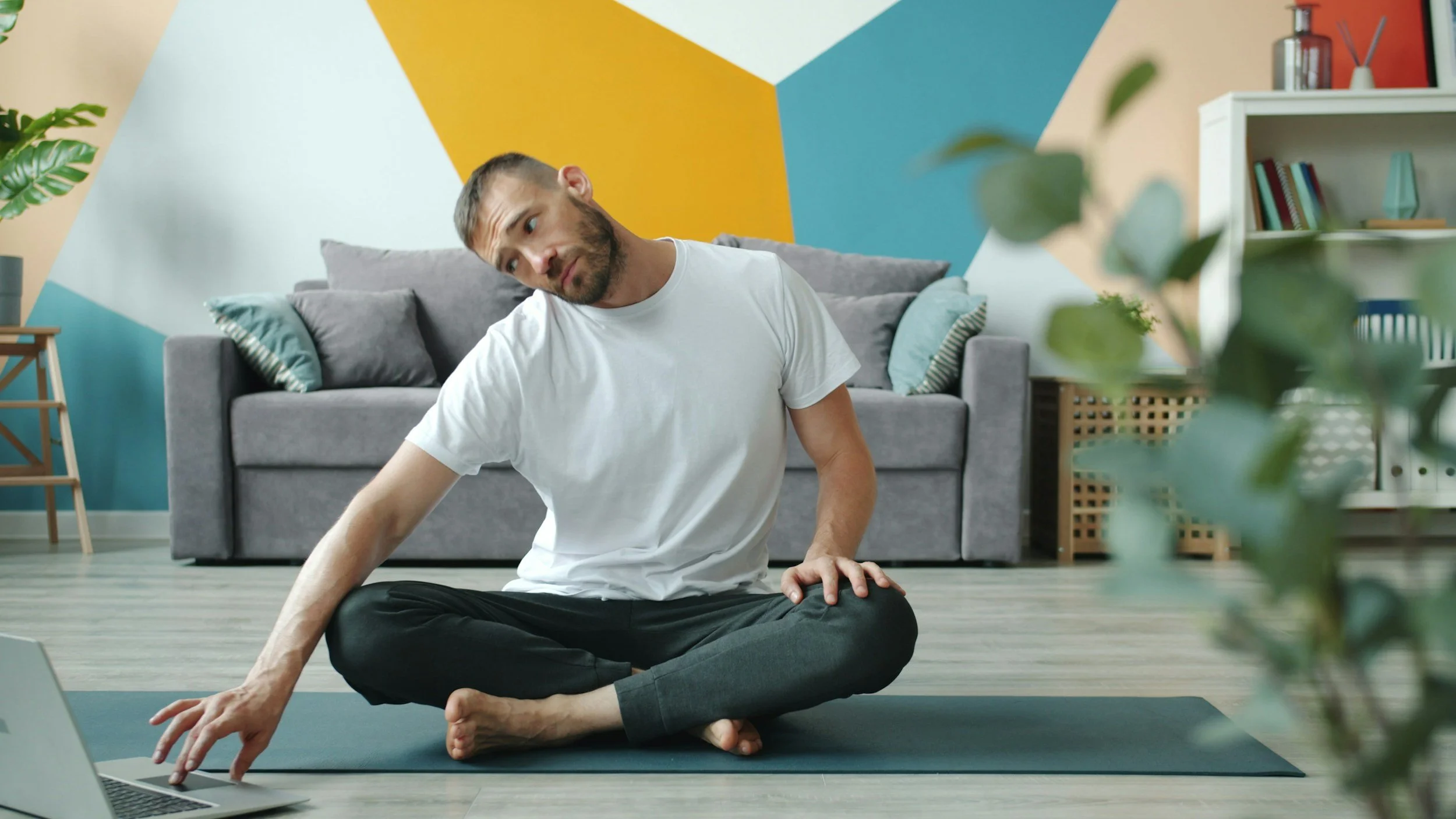 Man sitting cross-legged on yoga mat in living room, stretching with laptop nearby, colorful geometric wall background, indoor plants and furniture.