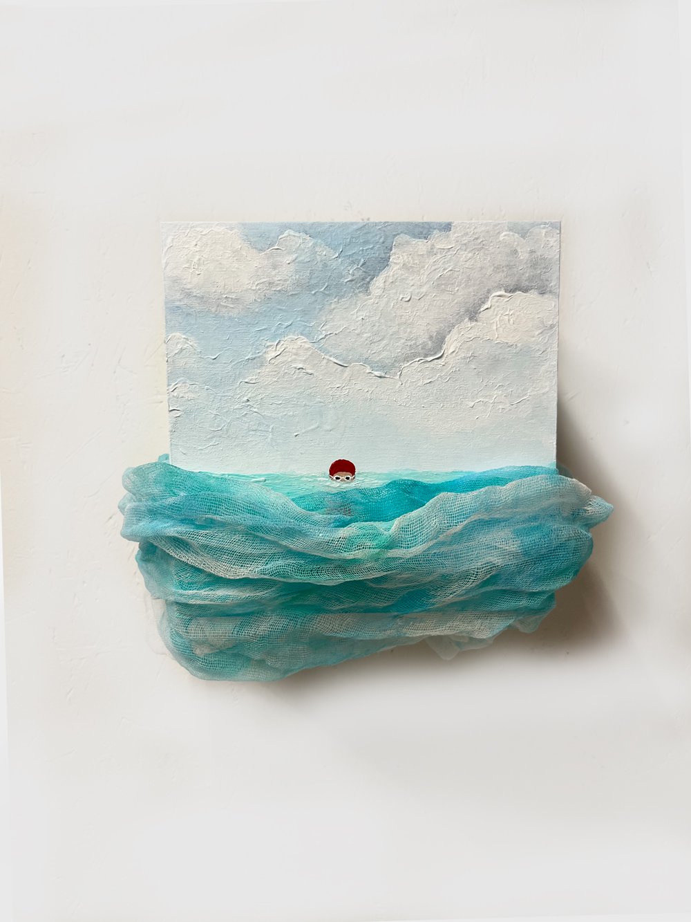 Mixed media art piece featuring a painted sky with clouds, ocean waves made of fabric, and a small figure with red hair and sunglasses in the water.