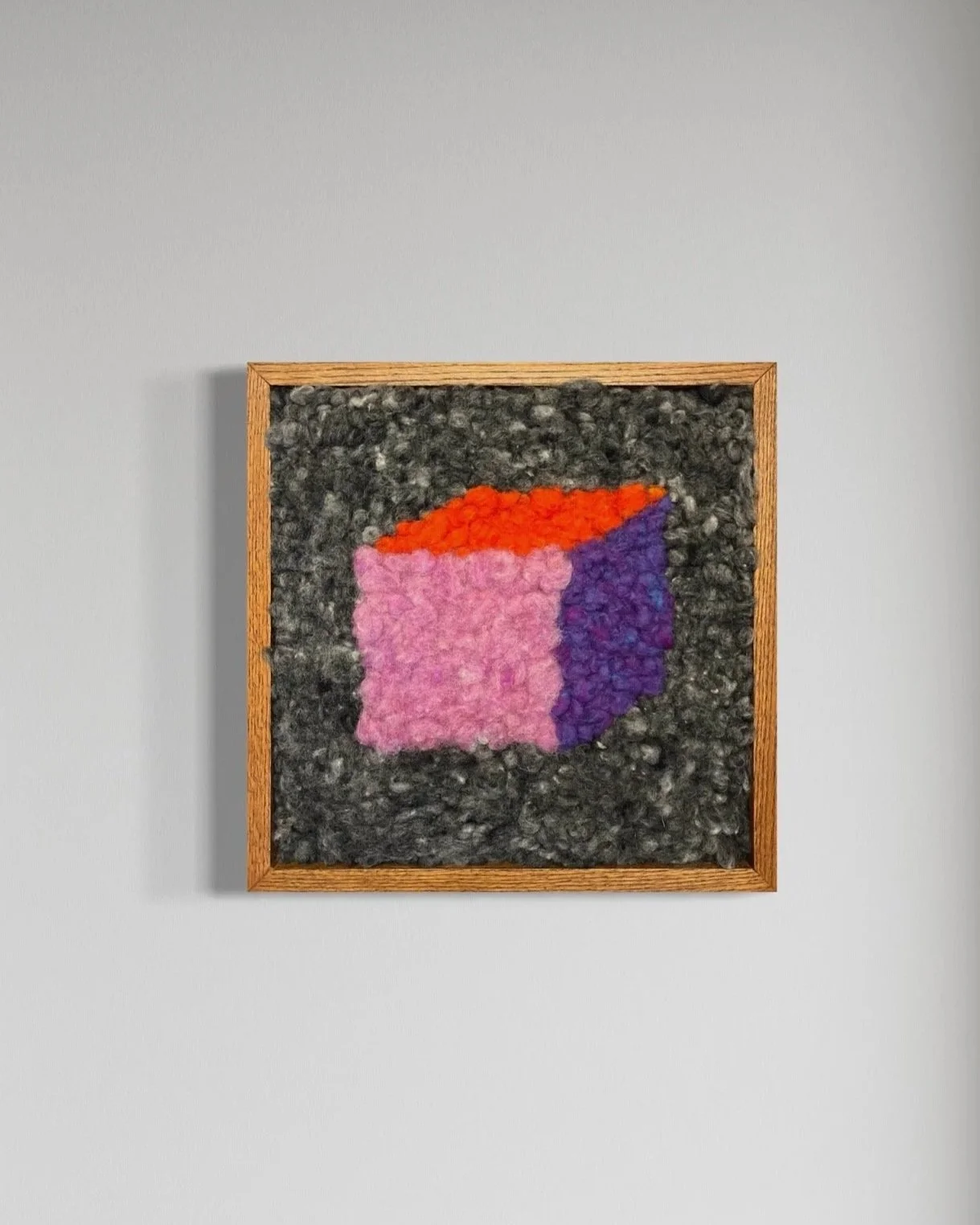 Textile art with a textured, multicolored abstract shape in pink, purple, and orange, framed in wood, hanging on a white wall.