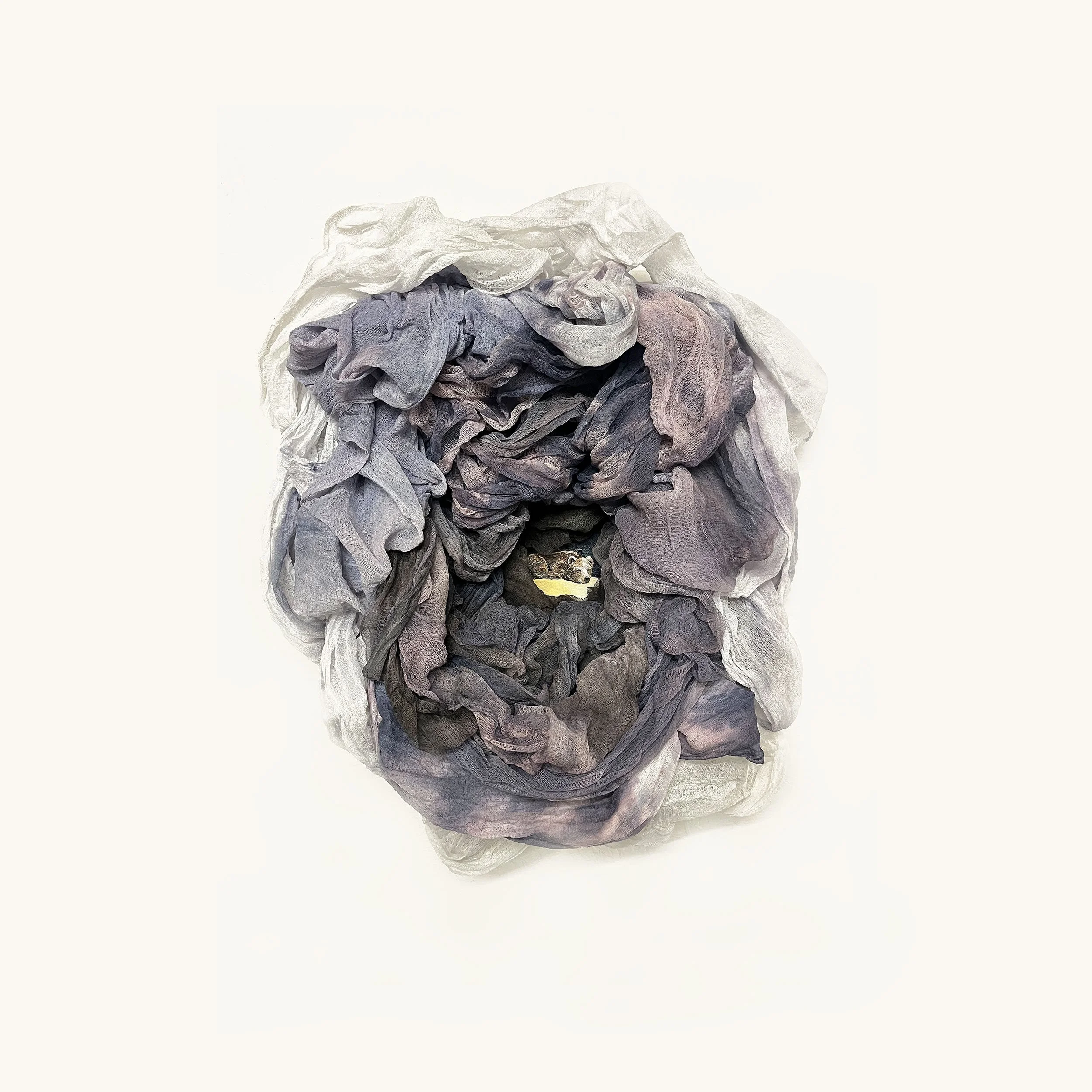 Pile of crumpled laundry or fabric in shades of purple, gray, and white with a small animal peeking through a hole in the center