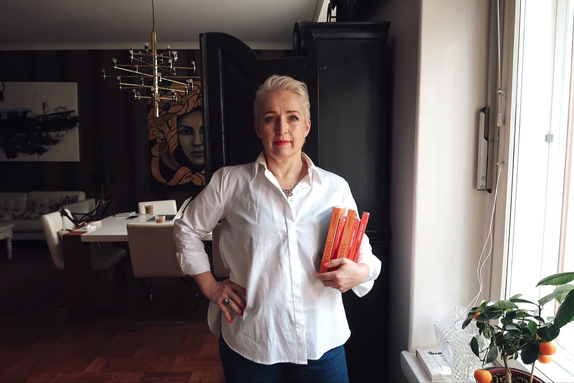 Nainen seisoo sisätiloissa, hänellä on lyhyet vaaleat hiukset ja hän pukeutuu valkoiseen paitaan ja sinisiin farkkuihin. Hän pitää kädessään kolme kirjaa ja seisoo ikkunan vieressä.