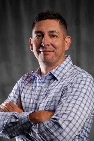 A man with short dark hair, wearing a checkered blue and white button-up shirt, standing with arms crossed in front of a dark gray background.