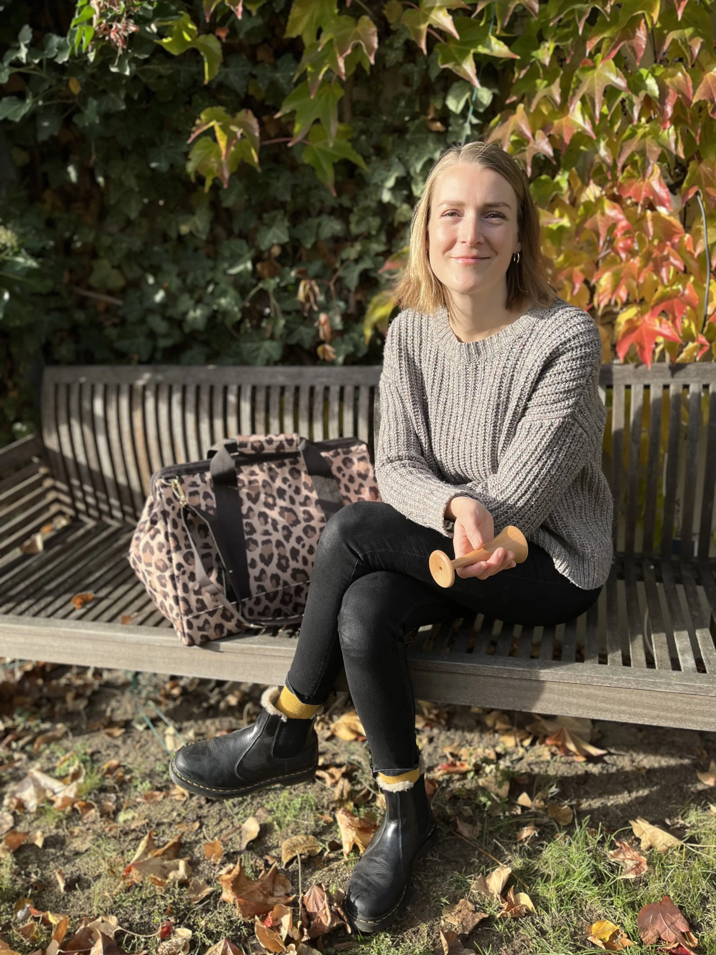 Junge Frau sitzt auf einer Gartenbank im Park, trägt eine graue Strickjacke, schwarze Jeans und schwarze Stiefel, hält eine Holzspielzeugrutsche in der Hand und hat eine Leopardentasche neben sich.