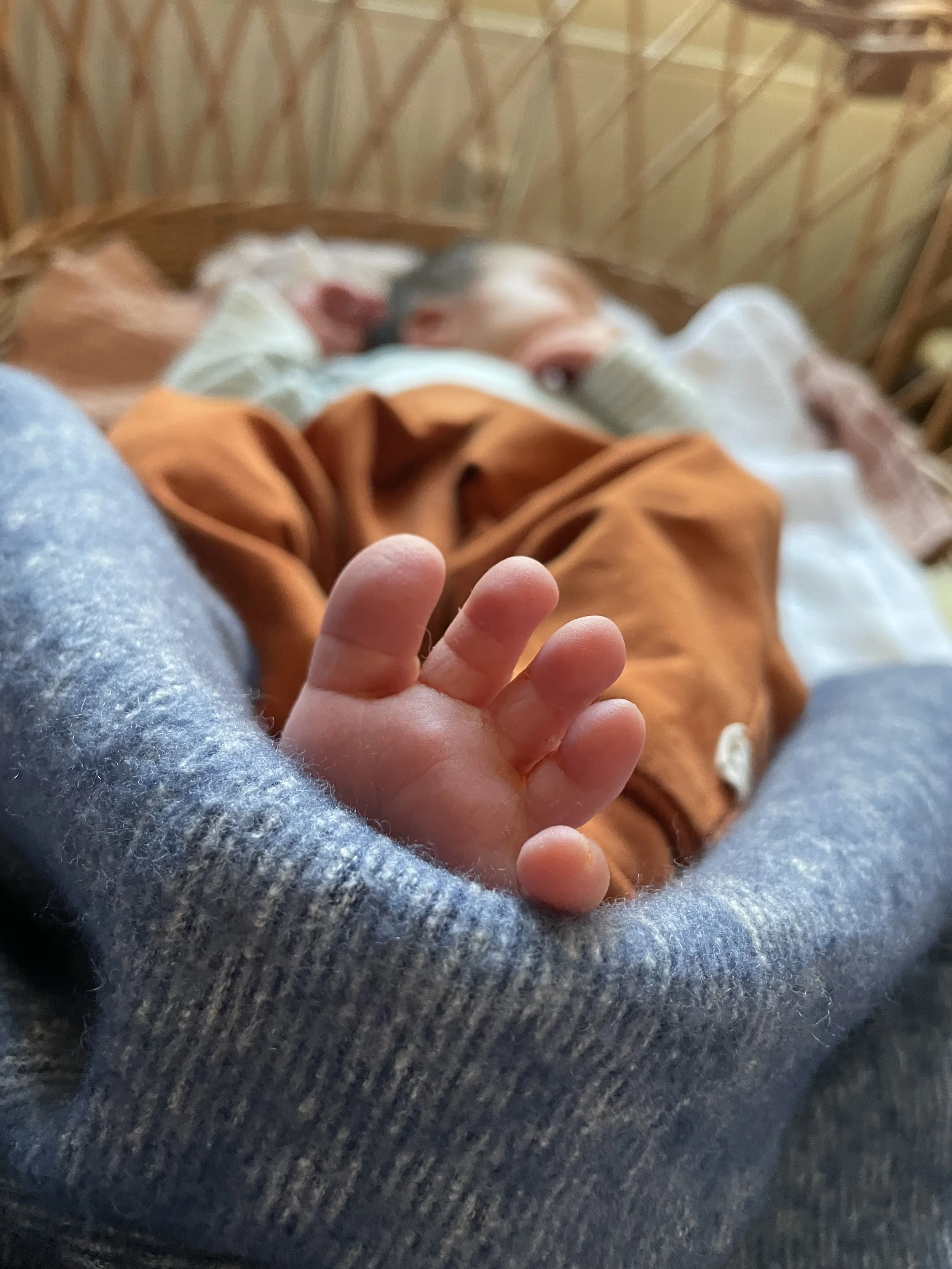 Ein kleines Baby liegt barfuß in einem Stubenwagen. Das Baby trägt eine orangefarbene Hose und ist teilweise von einer blauen Decke bedeckt. Im Vordergrund sind die nackten Füßchen.