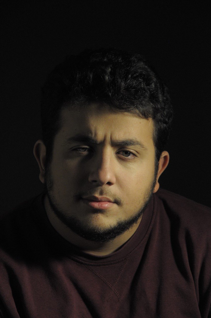 Portrait d'un homme avec des cheveux bouclés foncés, barbe, portant un sweat marron foncé, regardant vers l'objectif, fond noir.