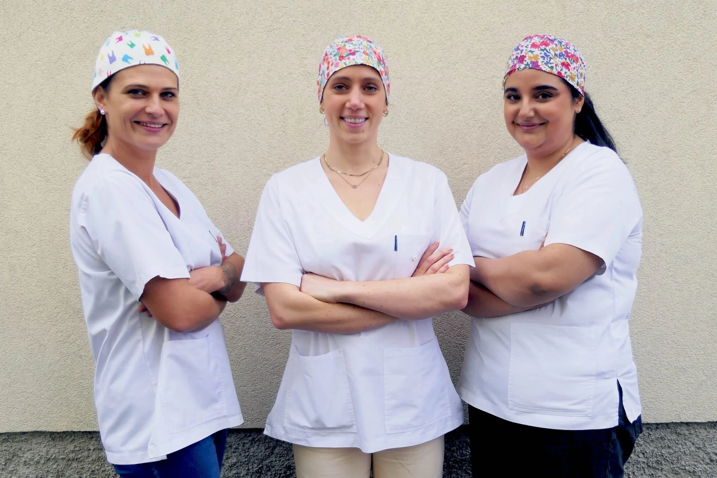 Trois femmes médecin ou infirmière souriantes portant des blouses blanches et des bonnets colorés, posant contre un mur beige.