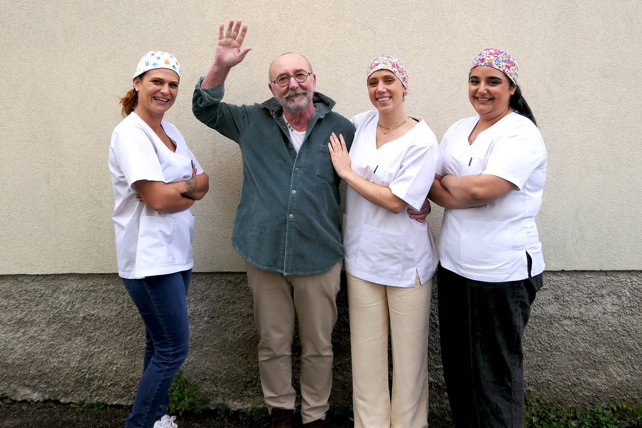 Quatre personnes, trois femmes en uniforme médical et un homme au centre, posent devant un mur beige, souriants. La femme à gauche a des cheveux roux et porte une blouse blanche, la femme à droite a des cheveux noirs et porte également une blouse blanche, la femme à droite porte une blouse blanche et un bandana floral, et l'homme au centre a une barbe grise et porte une chemise grise. La femme de droite pose sa main sur l'épaule de l'homme, qui fait signe avec sa main.