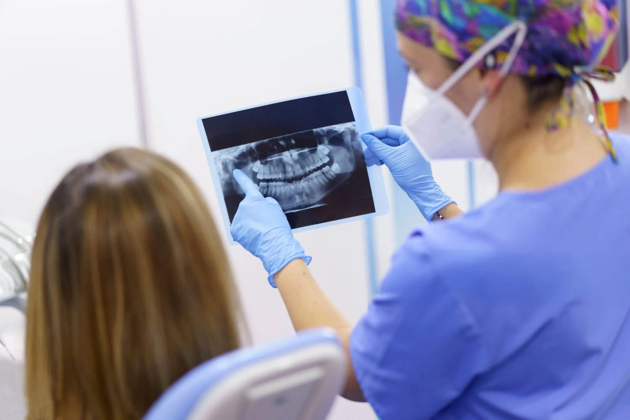 Une professionnelle de la santé examine une radiographie dentaire, portant une blouse bleue, un masque facial blanc, des gants bleus, et un chapeau coloré dans un cabinet médical lumineux.