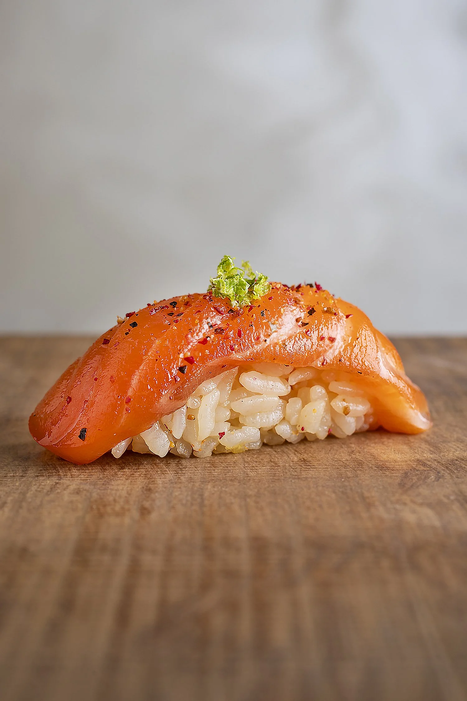 A piece of sushi with a slice of smoked salmon on top, garnished with black pepper and a small green herb, served on a wooden surface. 