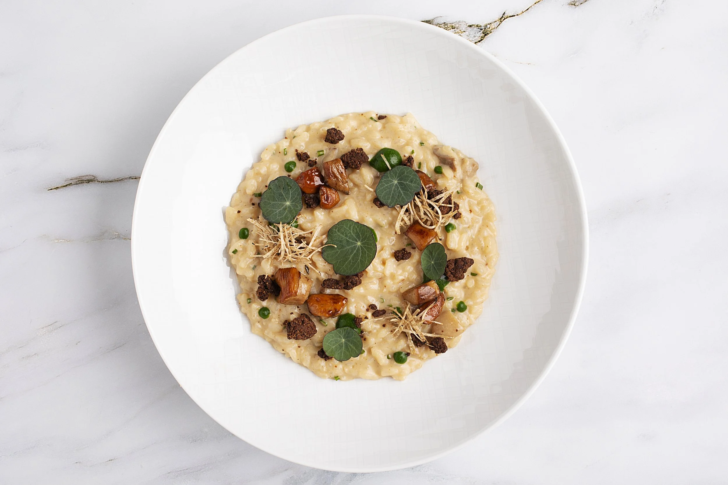 Creamy risotto on a white plate, garnished with green leaves, chopped nuts, diced vegetables, and small herb sprigs