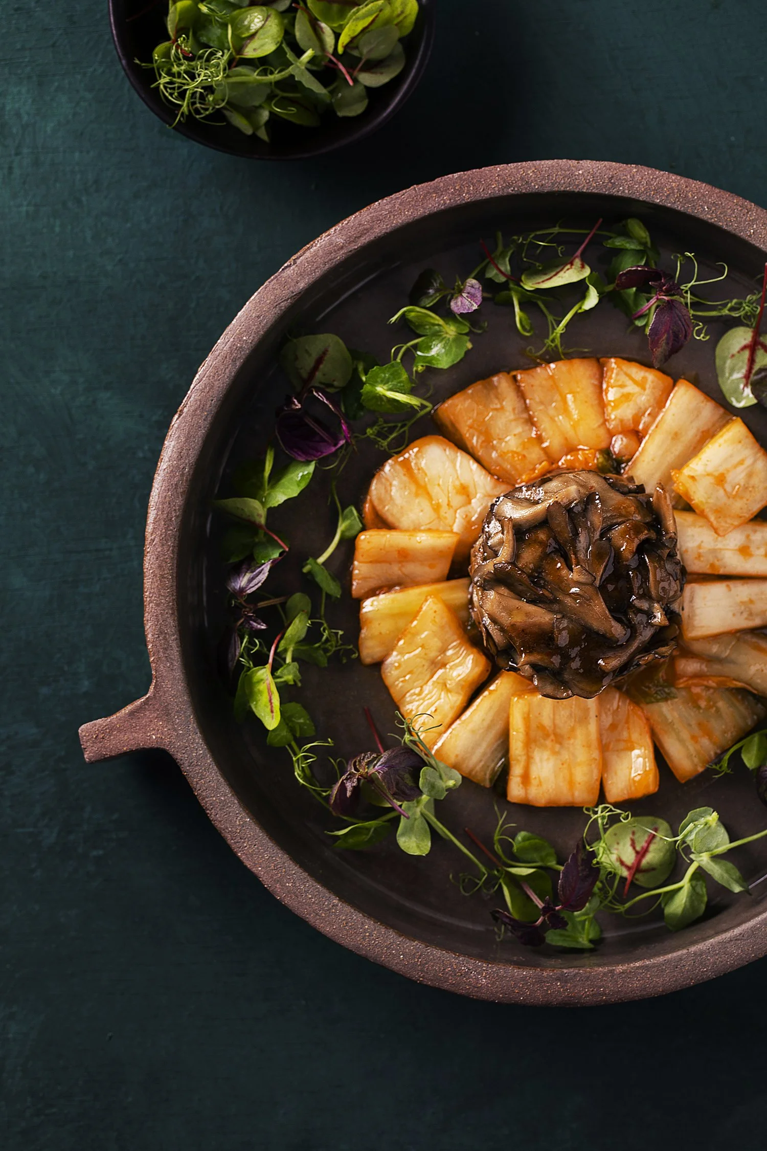 A top view of a black skillet with sliced radish arranged in a circular pattern, topped with a mound of sautéed mushrooms, and garnished with microgreens, placed on a dark green surface. 