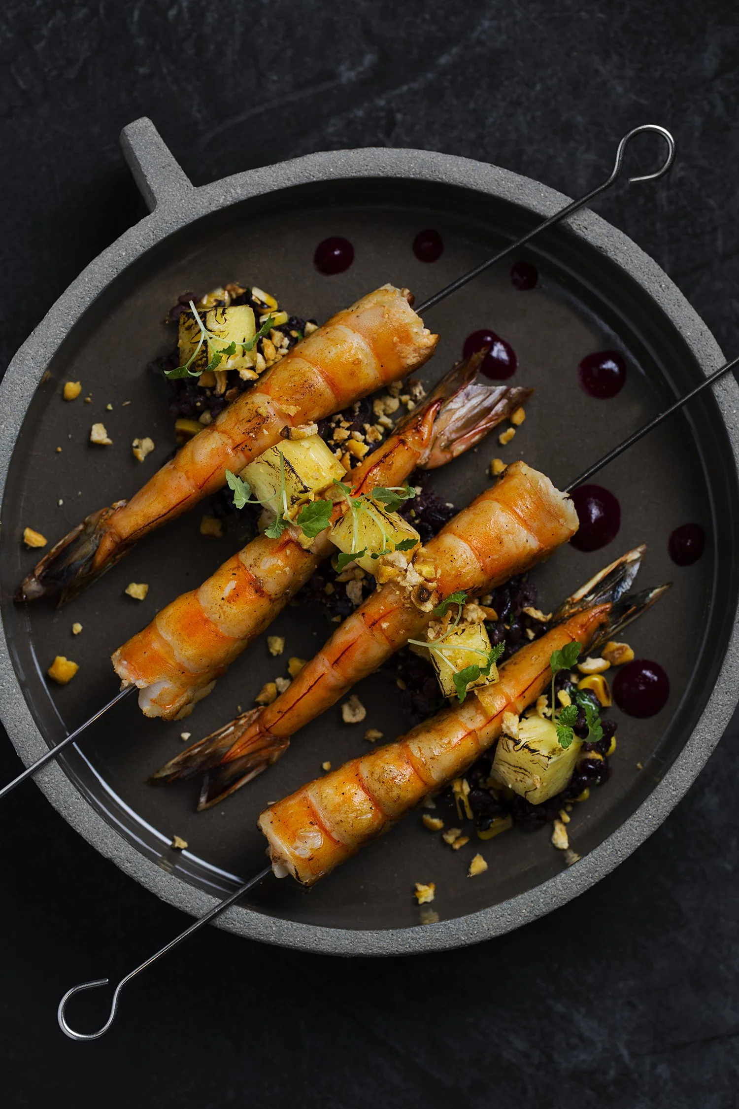 Grilled shrimp skewers garnished with herbs and lemon, served with chopped nuts and sauce dots on a black plate.
