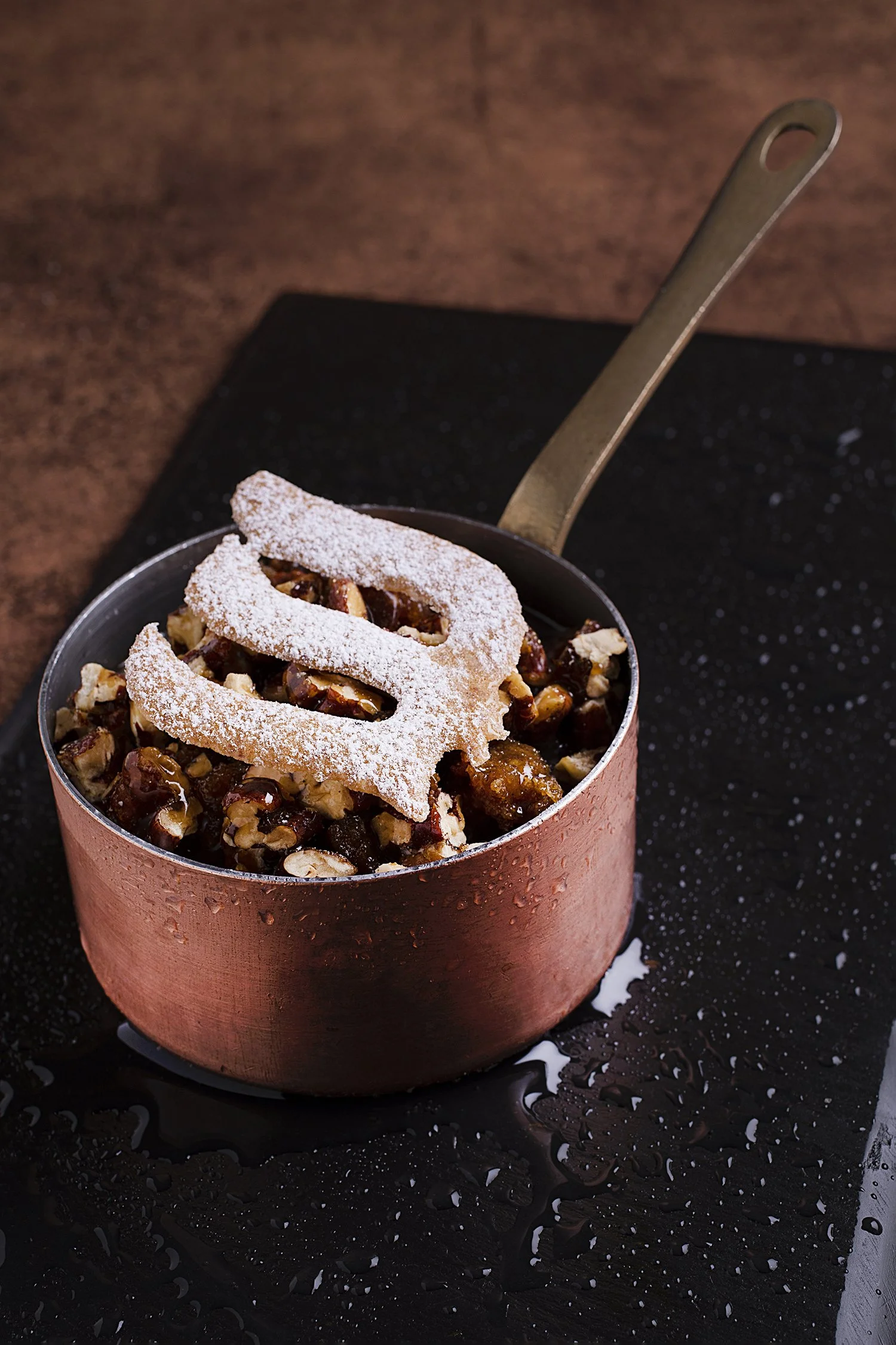 A small copper pot filled with a mixture of chopped nuts and dried fruits, topped with a dusting of powdered sugar and a decorative piece of puff pastry.