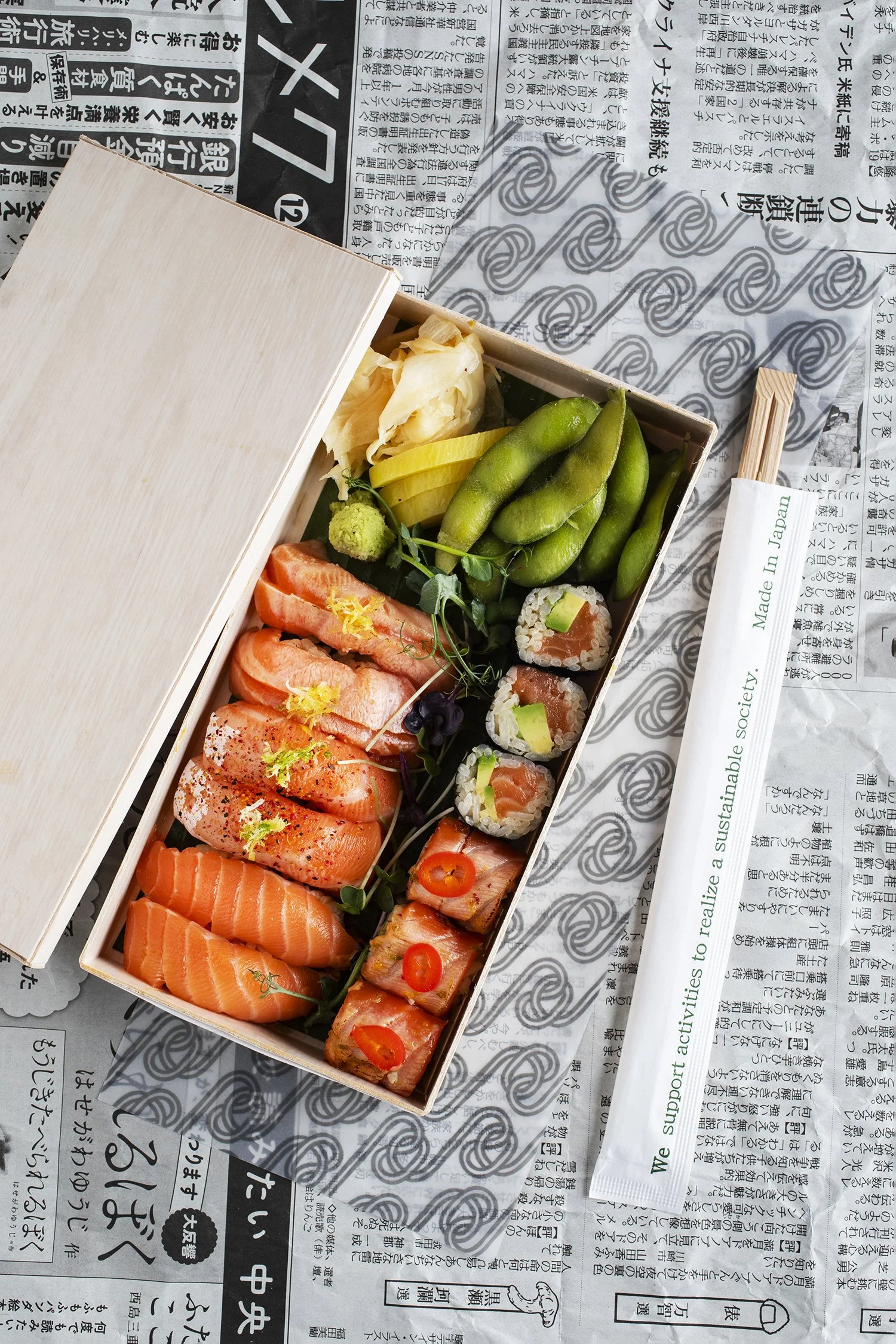 A wooden box of assorted sushi and Japanese side items on a newspaper-covered surface