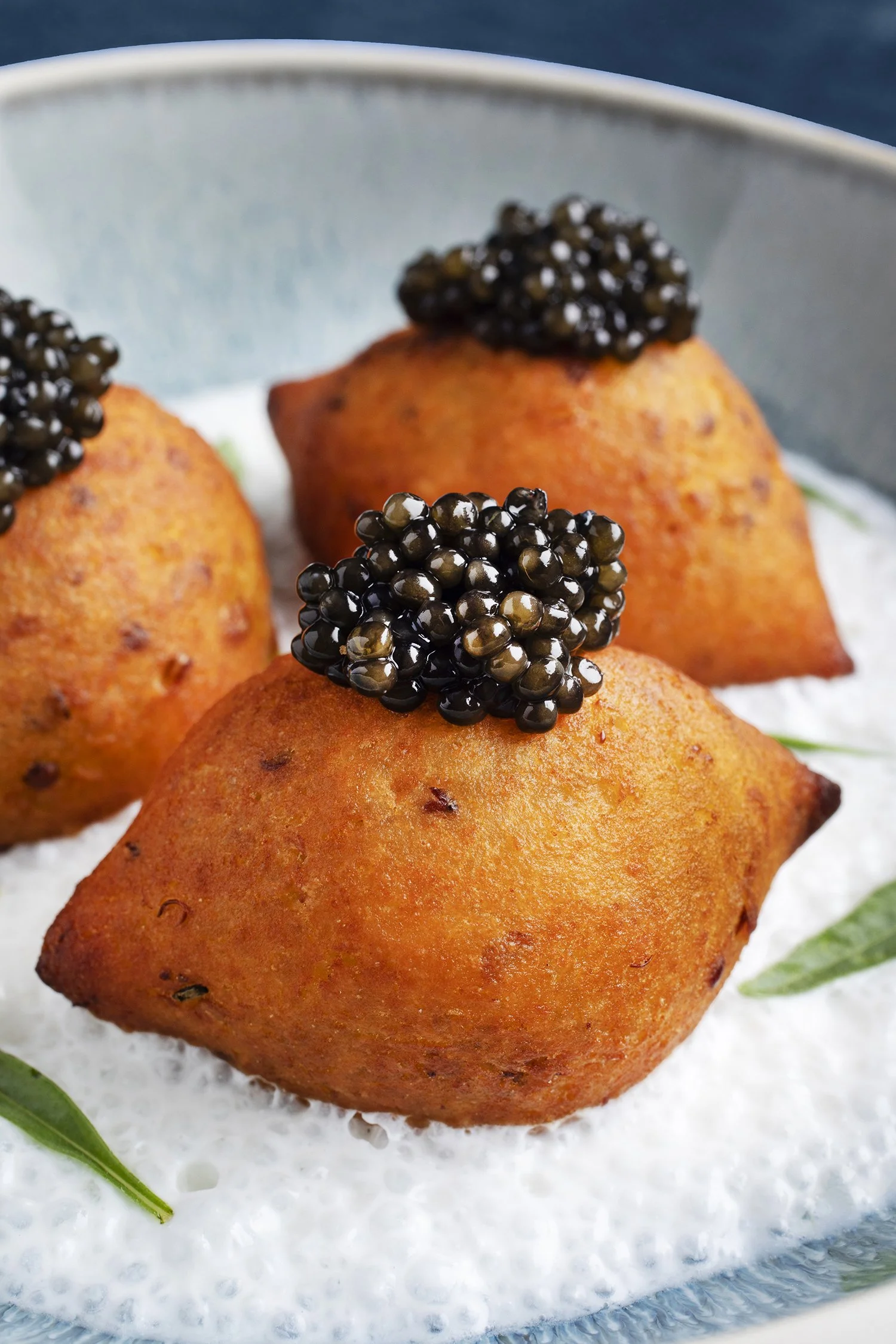Close-up of fried appetizers topped with black caviar, on a plate garnished with green leaves and a white sauce.
