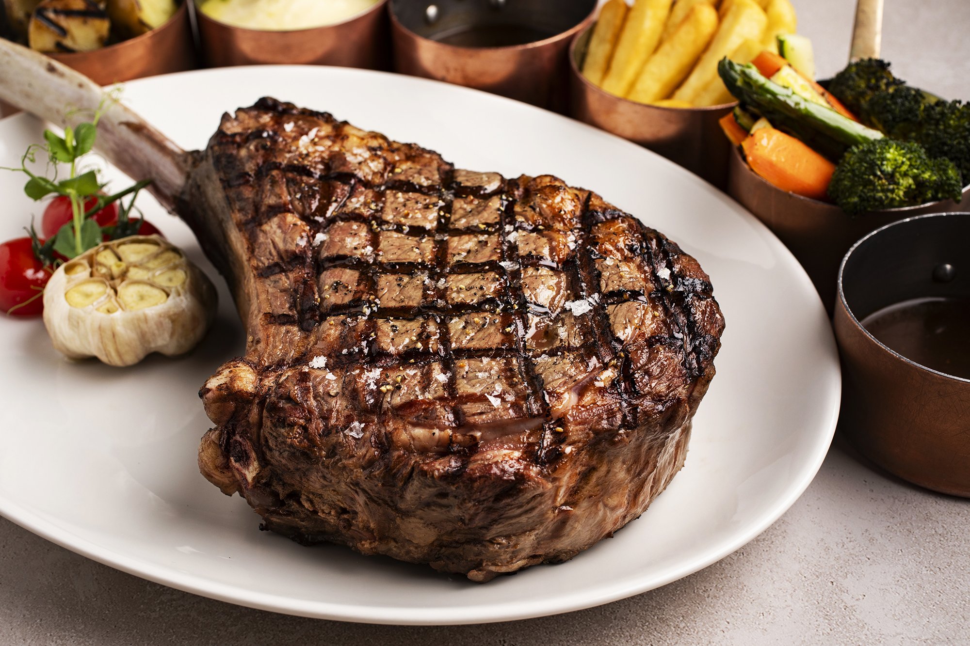 Grilled steak on a white plate with garlic, cherry tomatoes, and various side dishes including vegetables and fries. 