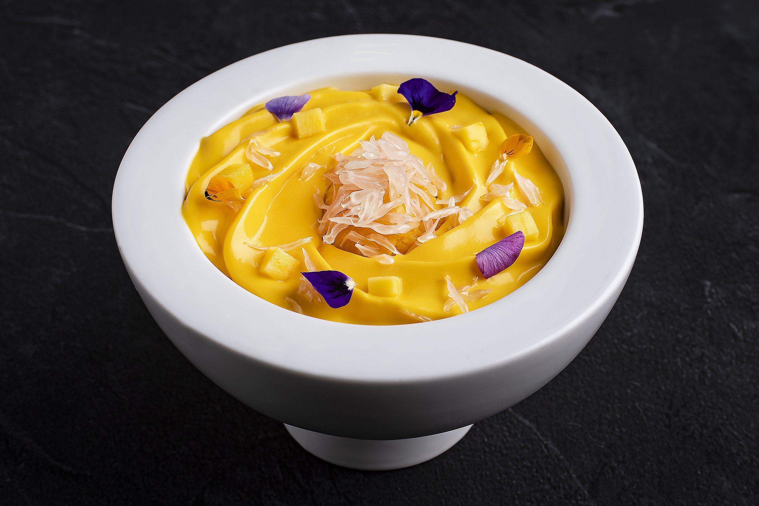 A white bowl with yellow mango pudding garnished with edible flower petals and small diced mango pieces, topped with shredded white fish or coconut.