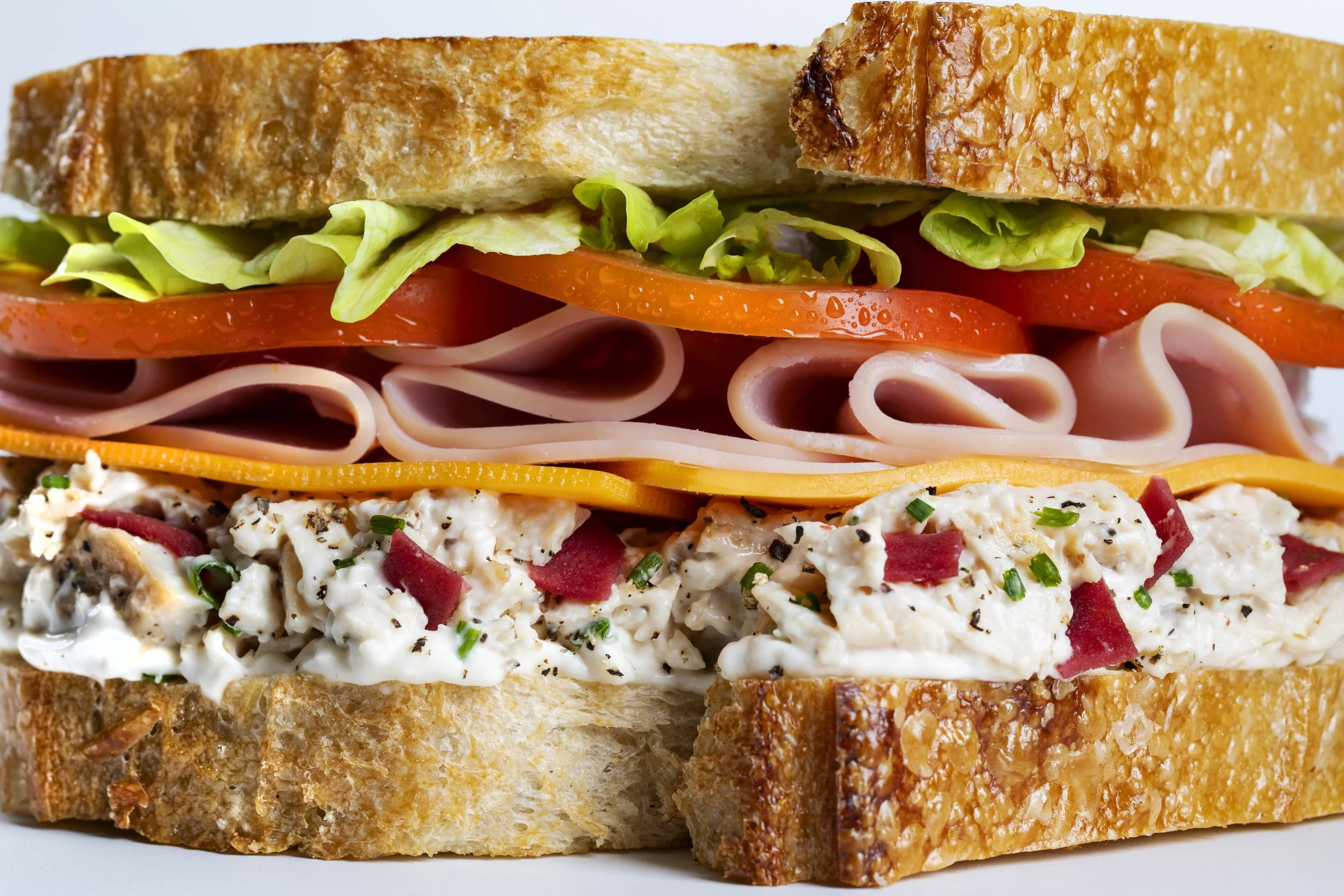 Close-up of a sandwich with toasted bread, lettuce, tomato slices, ham, cheddar cheese, chicken salad with red pepper pieces and green herbs.