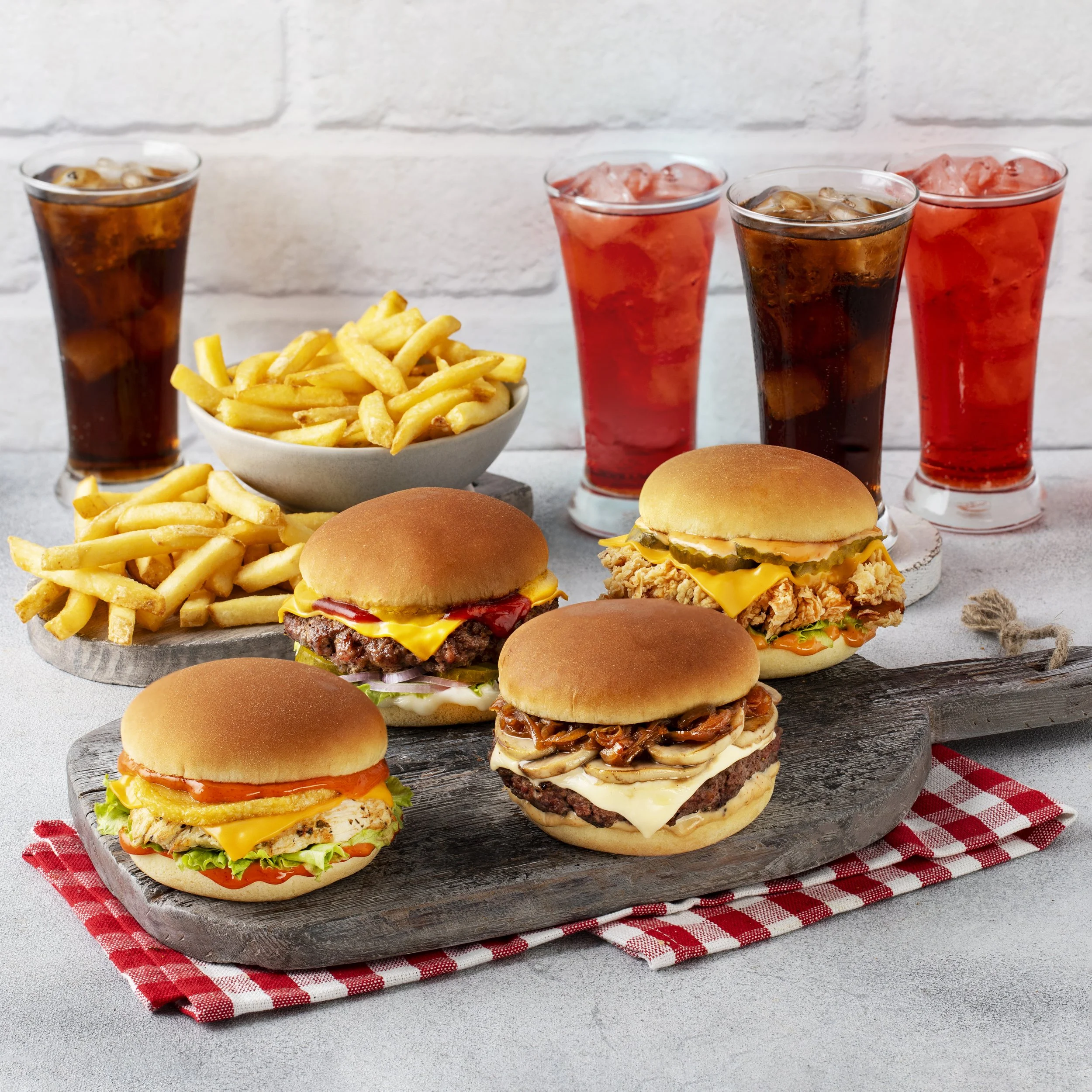 Four cheeseburgers with lettuce, pickles, and condiments, served on a rustic wooden platter with French fries and four glasses of soda