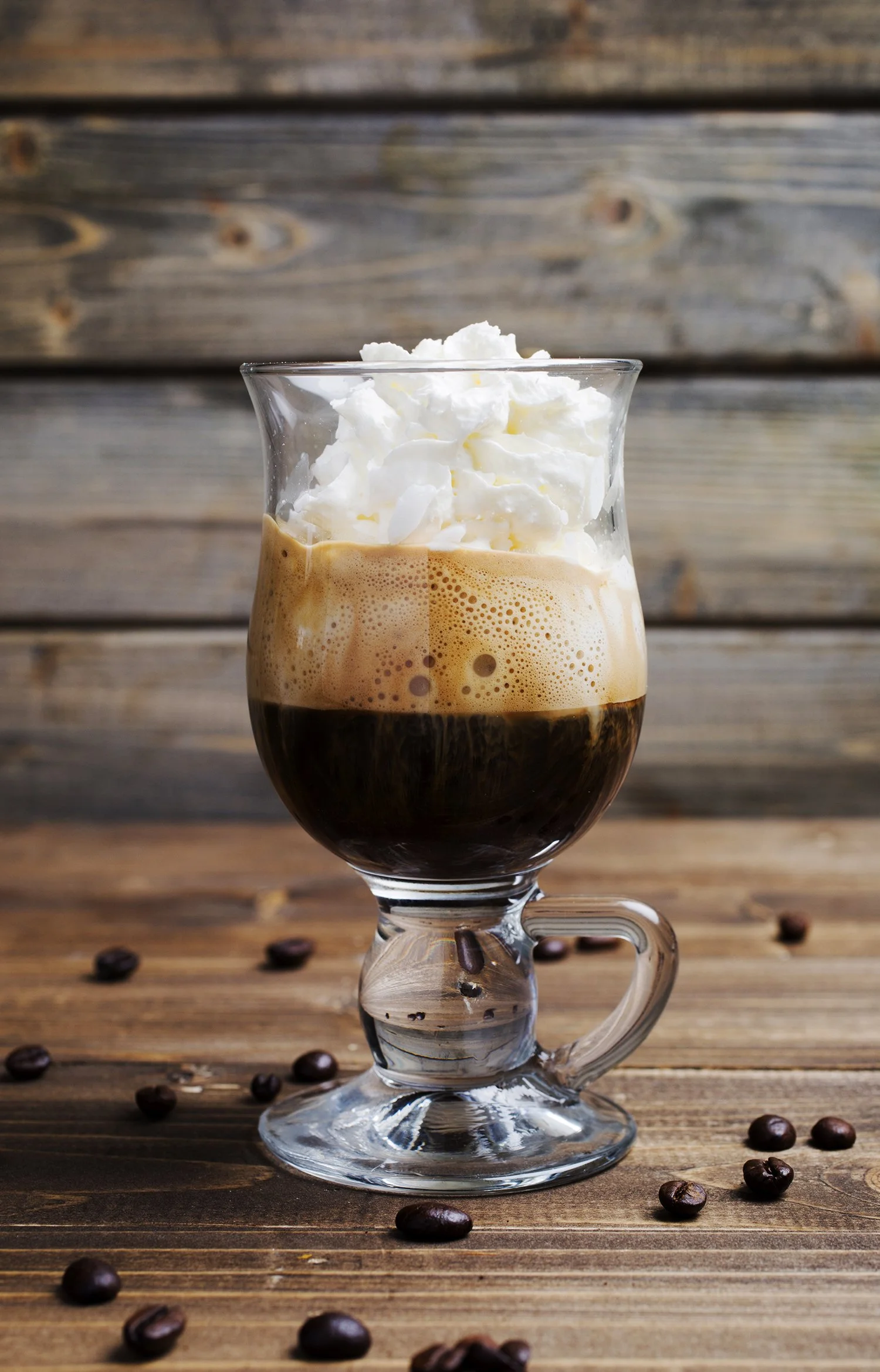 A glass of iced coffee topped with whipped cream and scattered coffee beans on a wooden surface. 