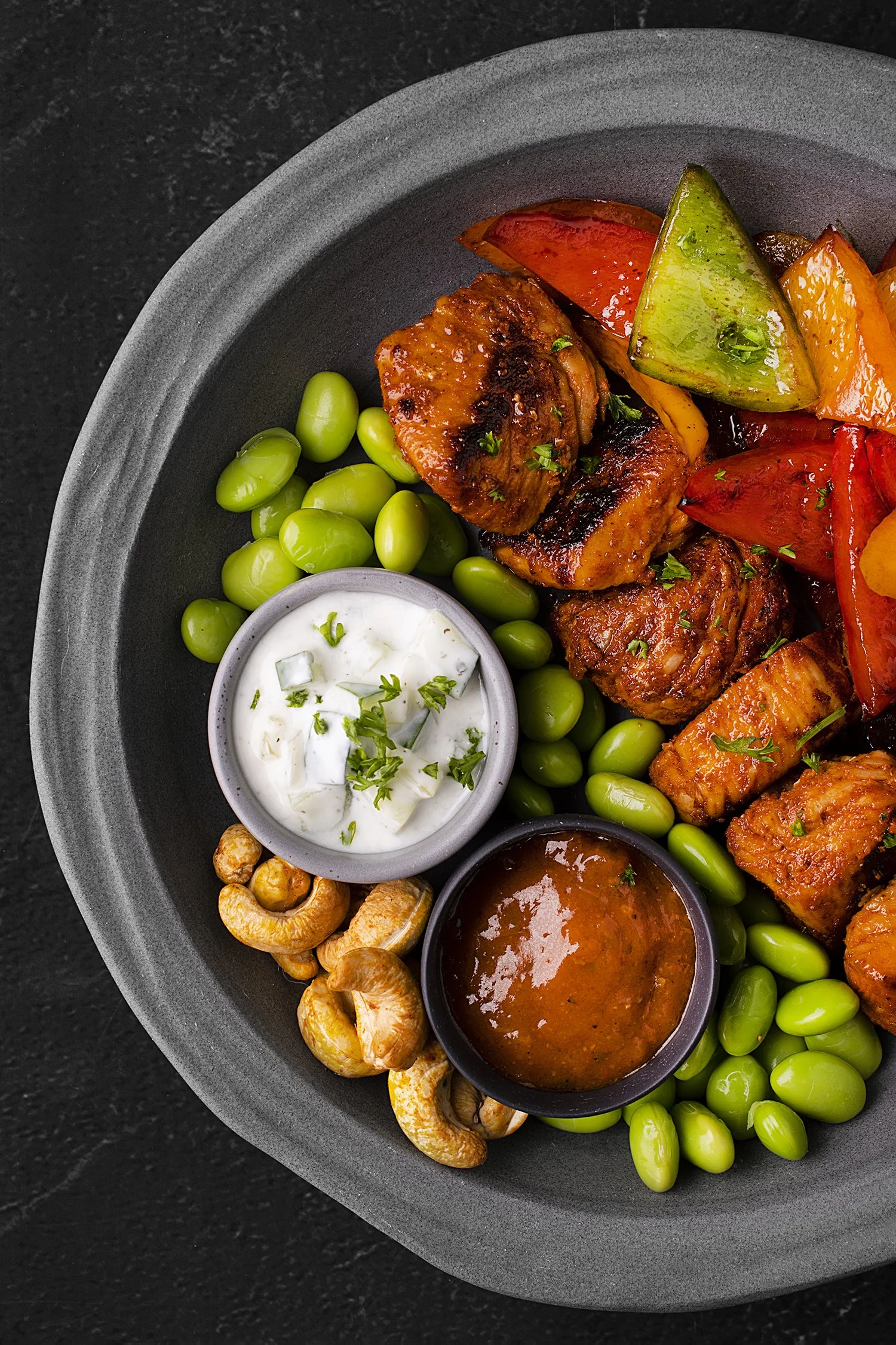 Grilled chicken with roasted bell peppers, green edamame, cashews, and two sauces on a black plate