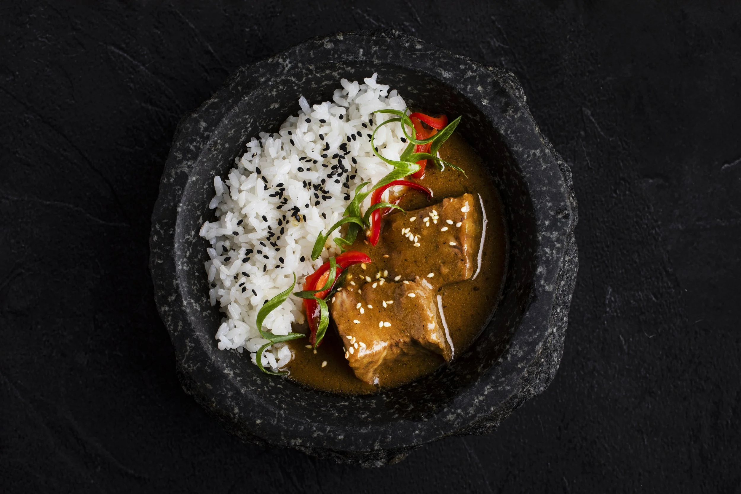A black stone bowl containing white rice topped with black sesame seeds, served with a portion of brown curry